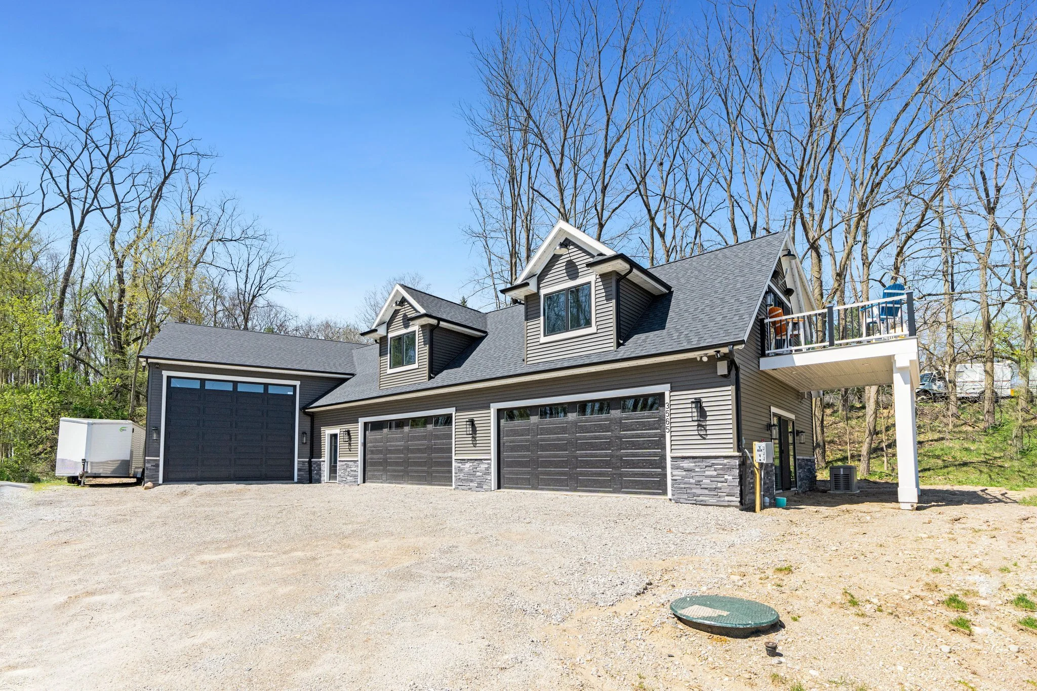 Modern house with gray siding, three garages, and a small balcony on the upper level, set in a wooded area with leafless trees and clear blue sky.