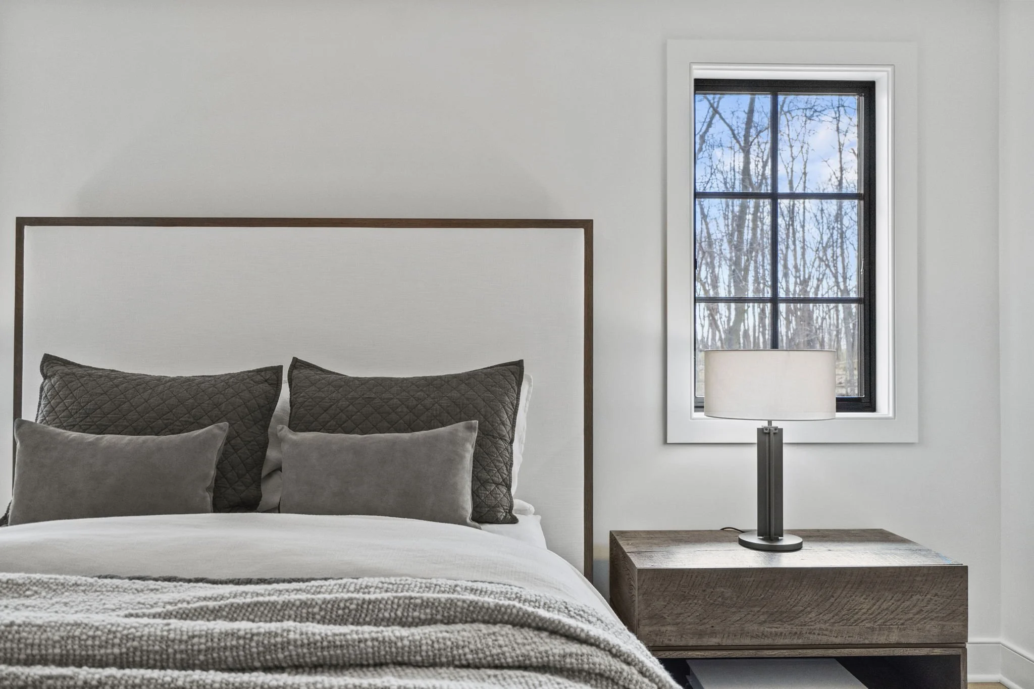 A neatly made bed with gray pillows, brown quilted pillows, and a textured blanket in a bedroom with a window showing trees outside, a wooden nightstand, and a table lamp.