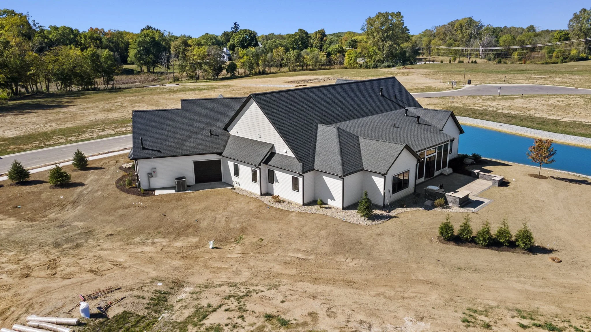 A newly built white house with a dark gray roof, surrounded by dirt yard, with a small tree and a backyard patio, overlooking a pond and a wooded area in the distance.