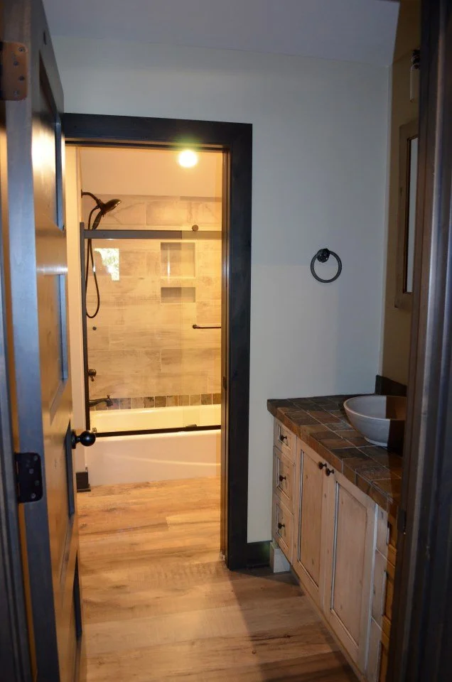 View into a bathroom showing a shower with a sliding glass door, a bathtub, and to the right, a wooden vanity with a tiled countertop and a vessel sink.