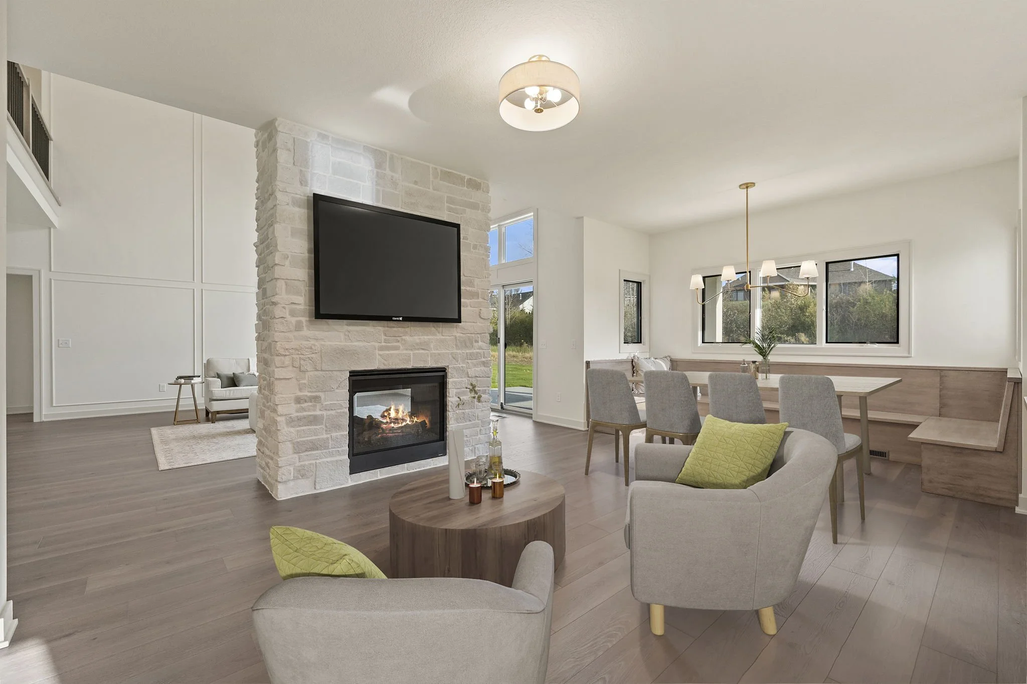 Modern living room with a fireplace, a large flat-screen TV, a round coffee table, armchairs with green pillows, a dining area with a wooden table and light gray chairs, and large windows overlooking a green outdoor space.