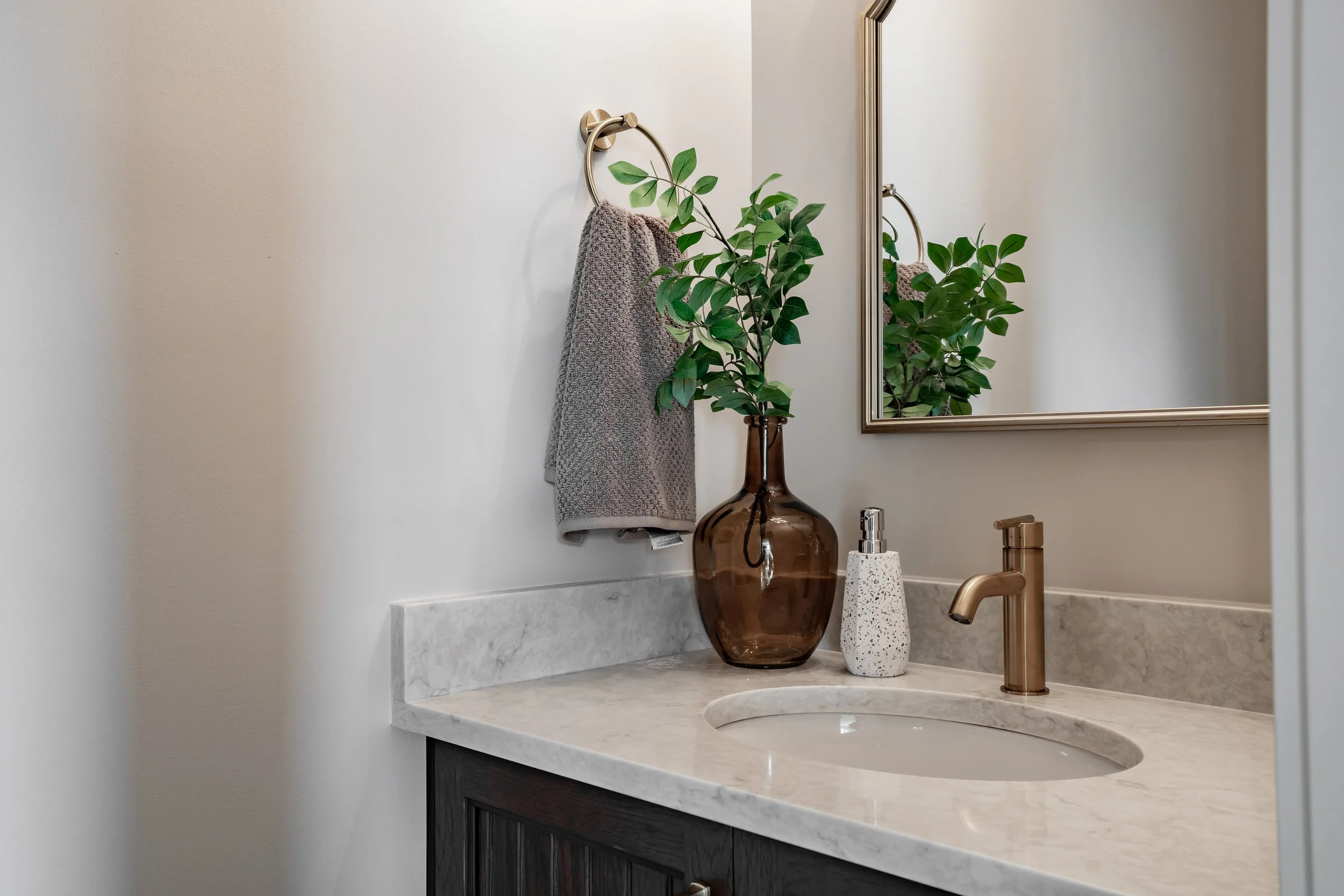 Bathroom vanity with marble countertop, brown vase with green leaves, soap dispenser, mirror, and gray towel.