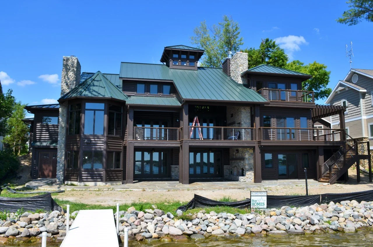 Large modern house with multiple levels and green metal roof, situated near a body of water, surrounded by rocks and trees, under a blue sky.