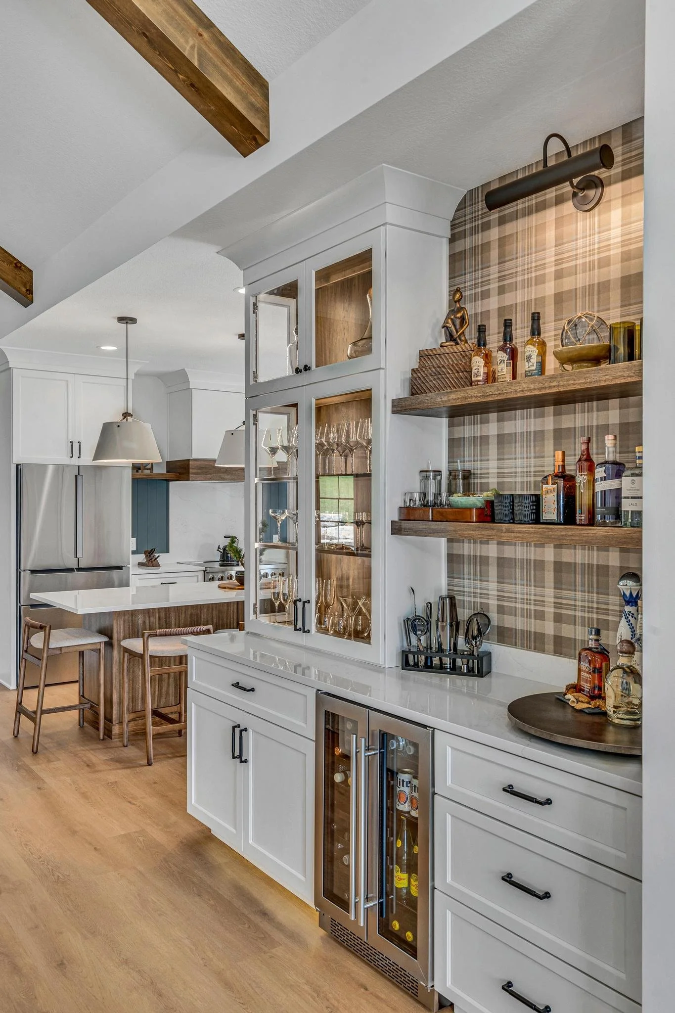 Kitchen with white cabinetry, open wooden shelves, glass display cabinet with glasses, wine fridge, and a countertop with liquor bottles and bar accessories, wood flooring, and part of a dining area with bar stools.