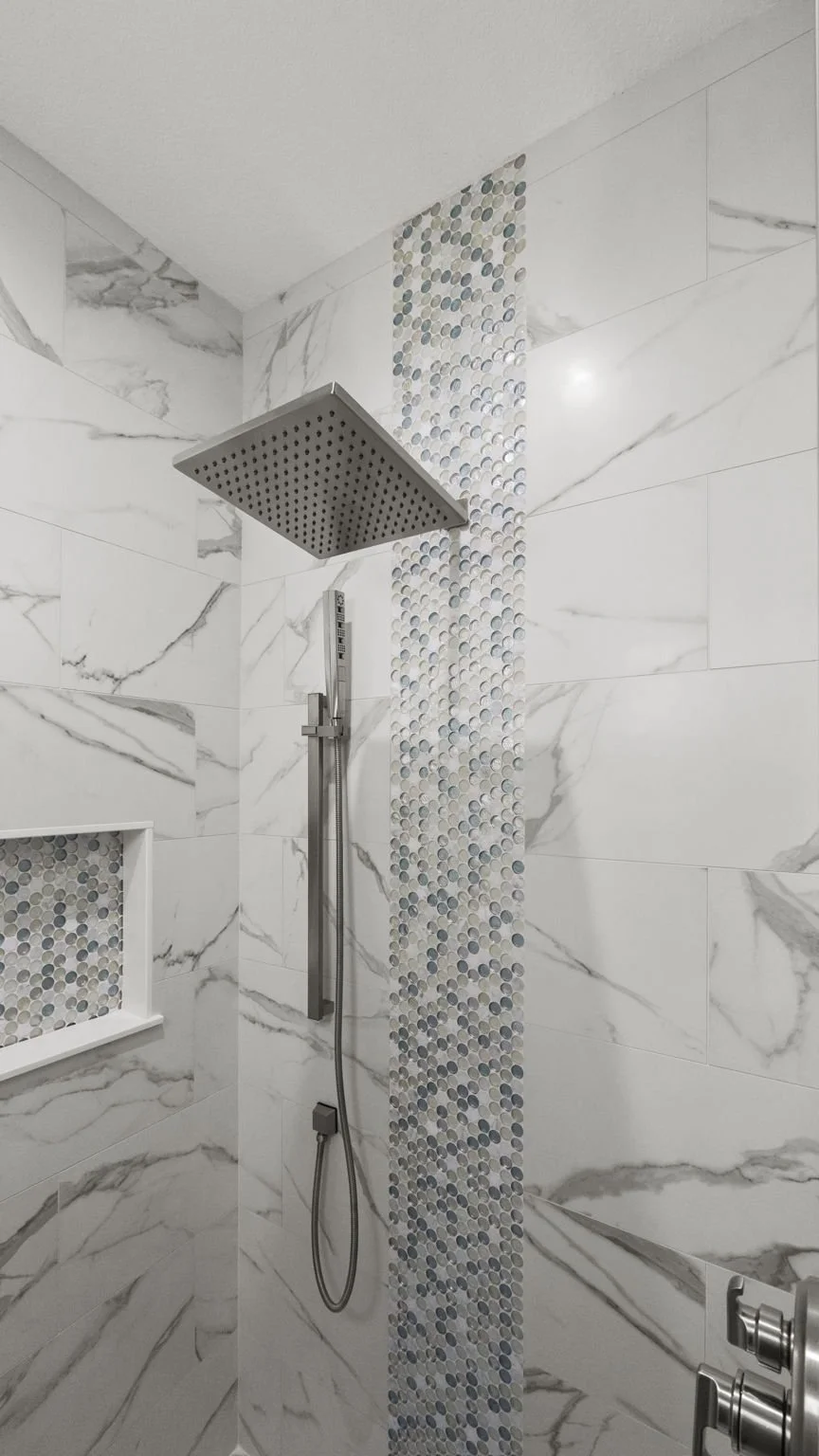 Modern shower with a large square rain shower head, marble tile walls, and a vertical strip of small, round, multi-colored mosaic tiles.