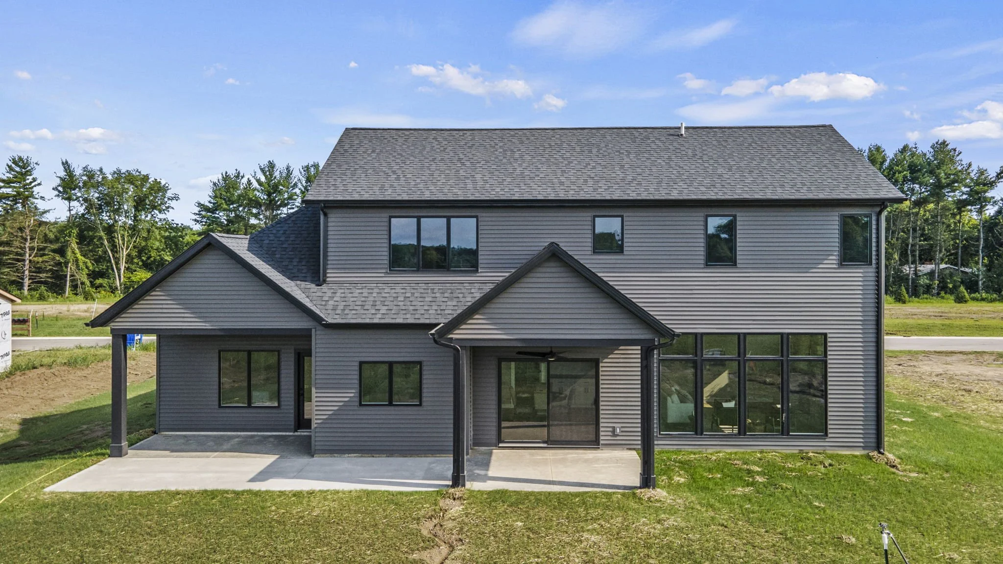 New gray two-story house with black window frames, a small covered porch, and a concrete patio in a grassy yard, surrounded by trees under a blue sky with clouds.