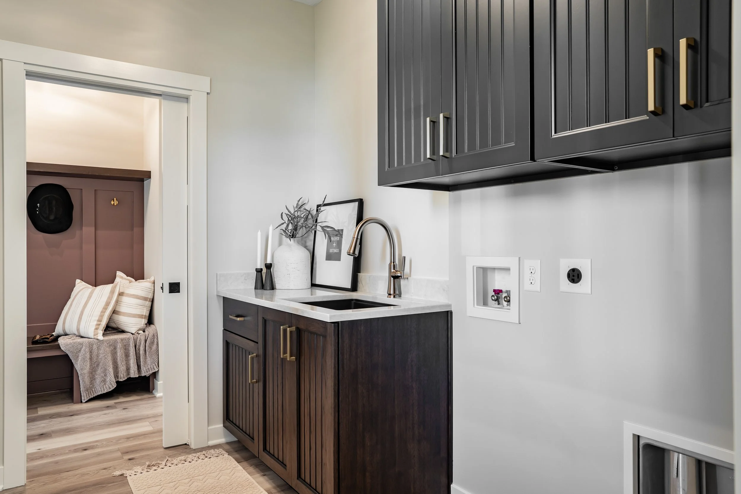 Small kitchen area with a dark wood cabinet under a white marble countertop, a small black sink, a chrome faucet, and decor including a white vase with branches, photo frame, and black and white candlesticks. Upper dark gray cabinets with brass handl