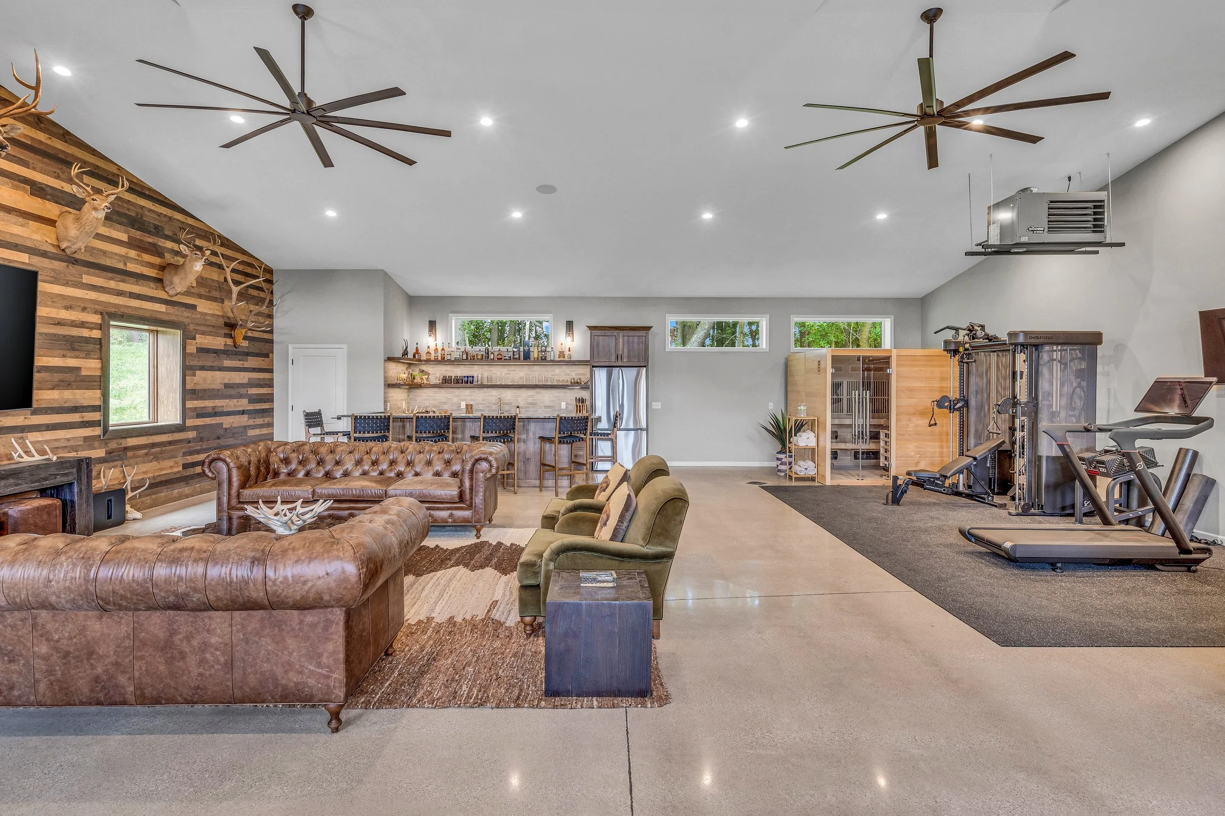 Spacious game room with bar area, leather couches, gym equipment, and deer head mounts on a wood-paneled wall.