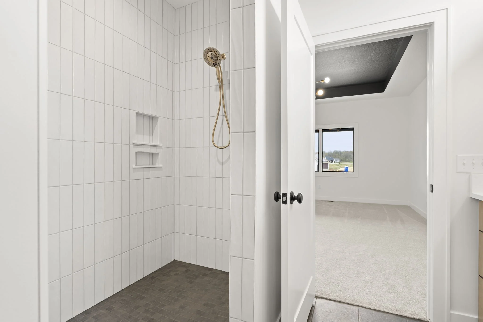 Empty shower with white tiled walls, two built-in shelves, a gold showerhead, and a gold hose, opening into a room with white walls, a window, and beige carpet.