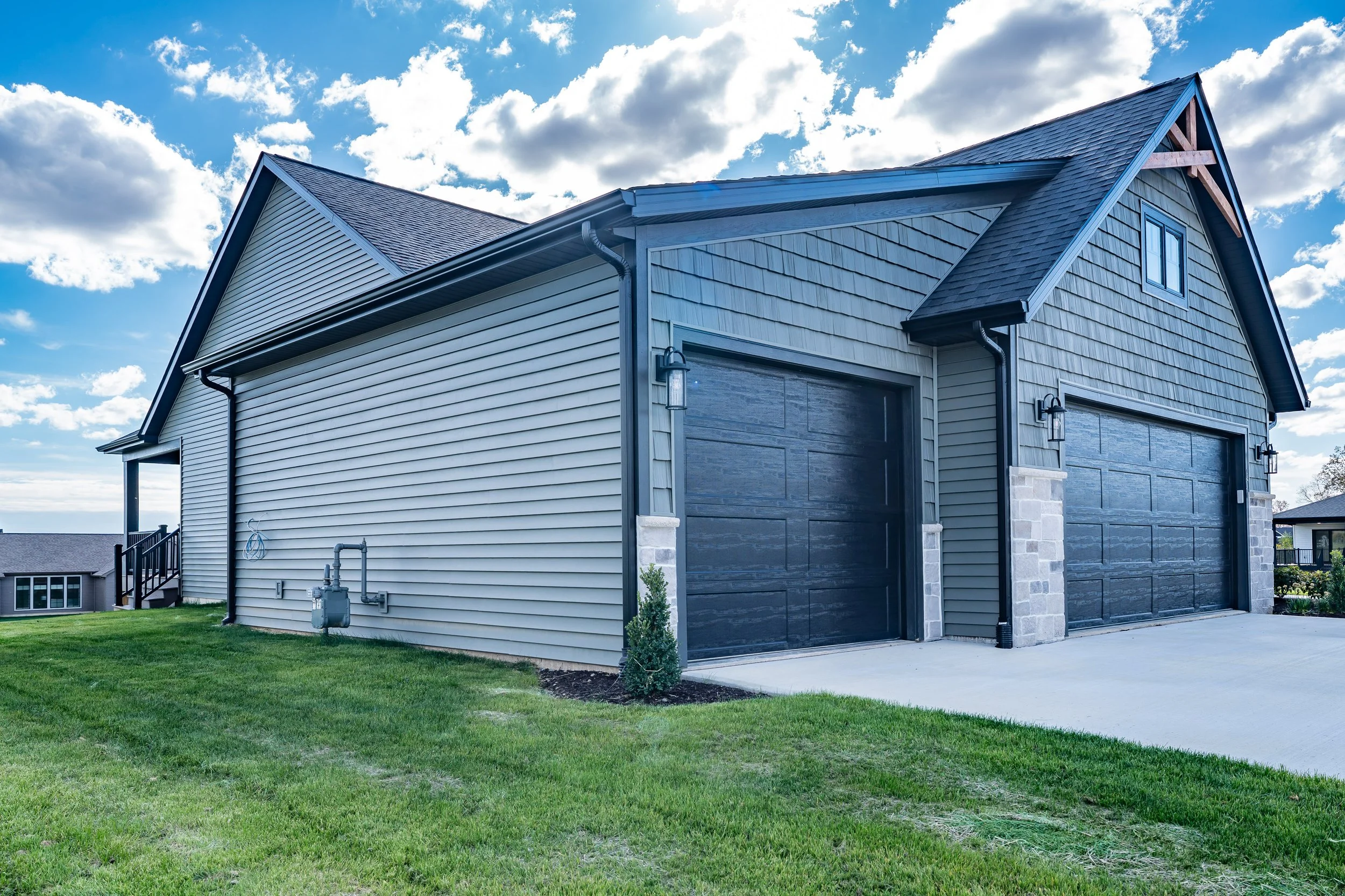 Exterior view of a modern house with a two-car garage, green grass lawn, and blue sky with scattered white clouds.