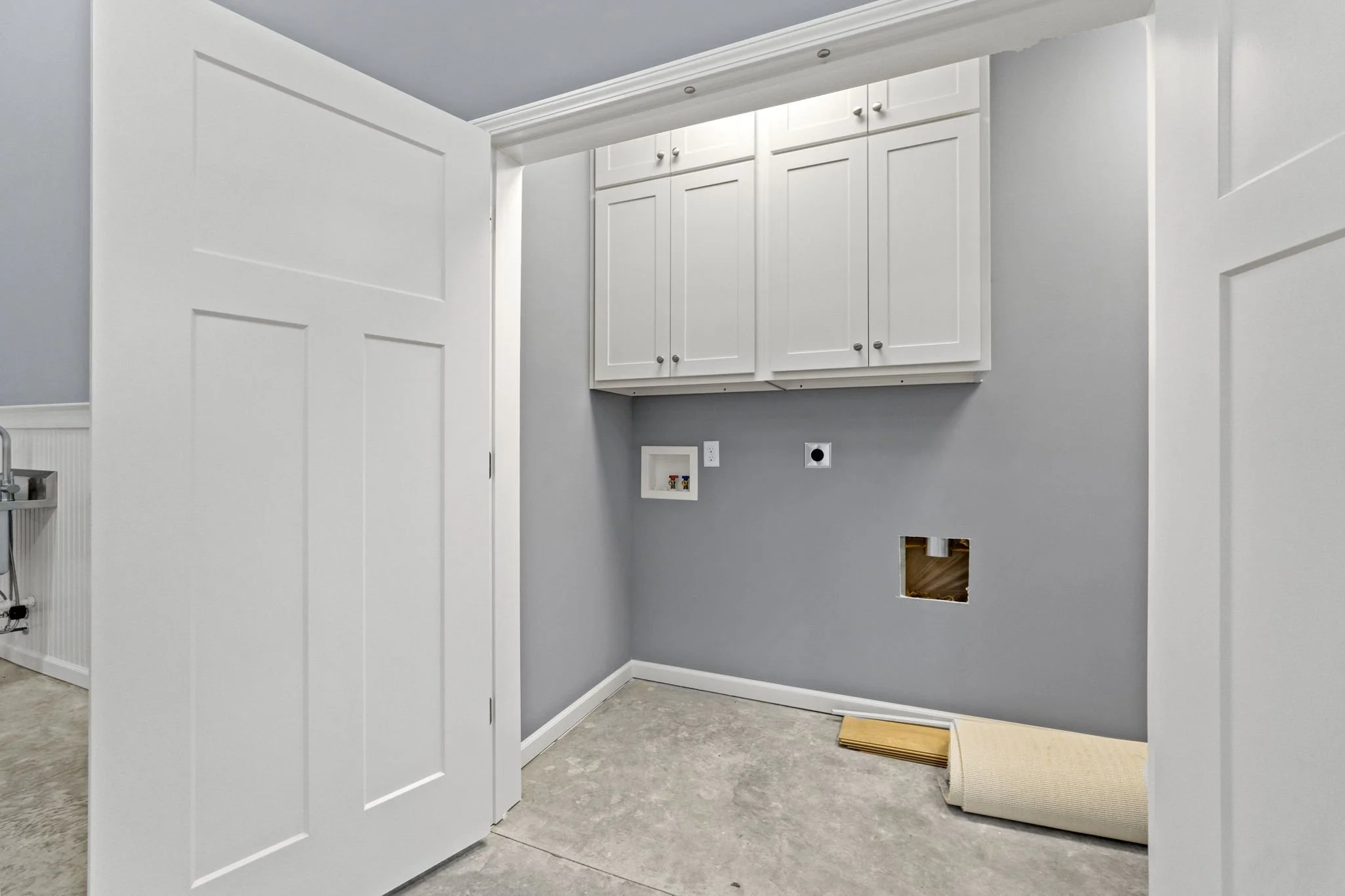 Laundering area with white cabinets, gray walls, and an empty space with plumbing hookups for a washer and dryer. A rolled-up carpet is on the floor.