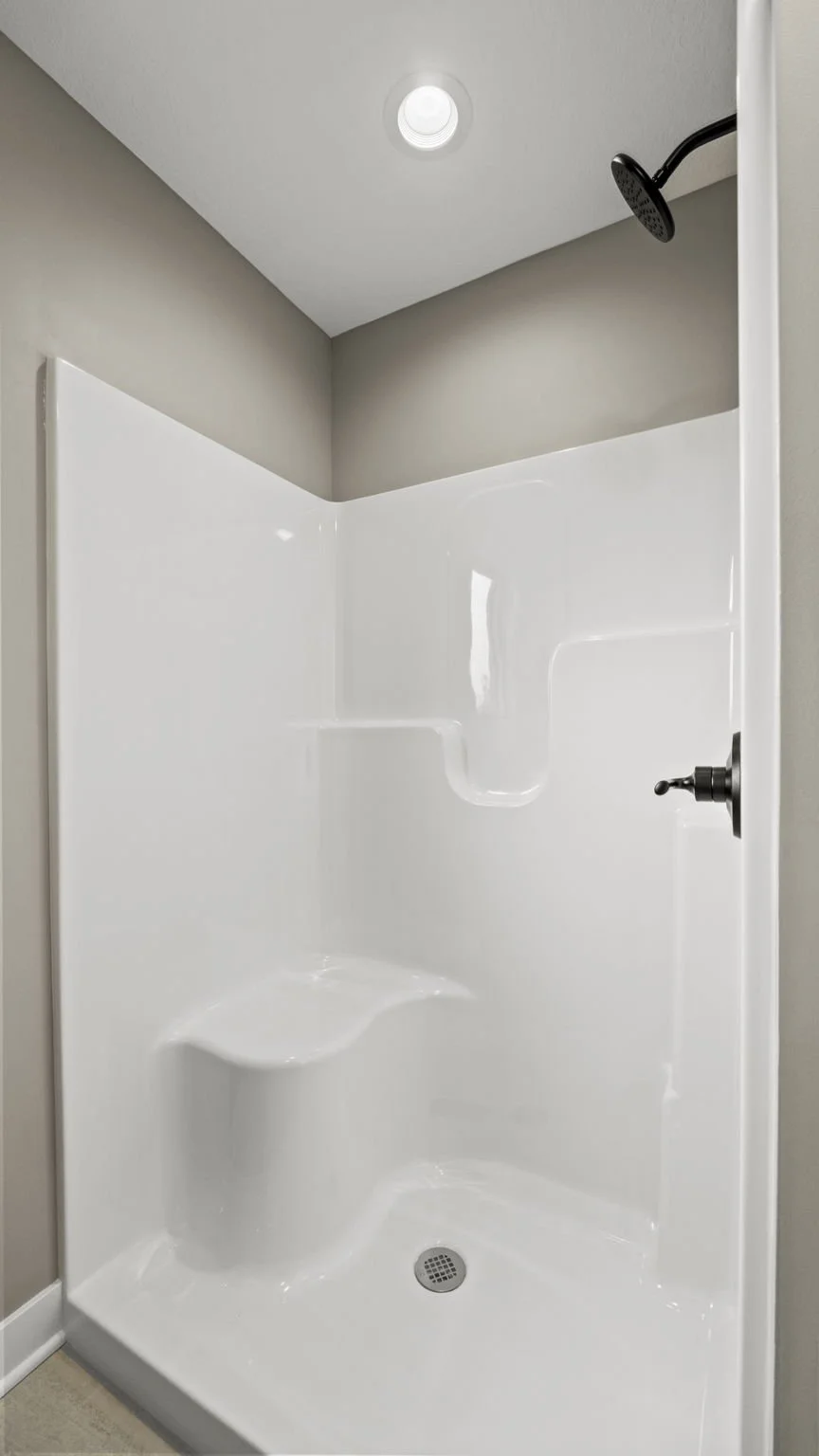Empty white shower stall with a built-in seat, a black showerhead, a ceiling light, and a drain on the floor.
