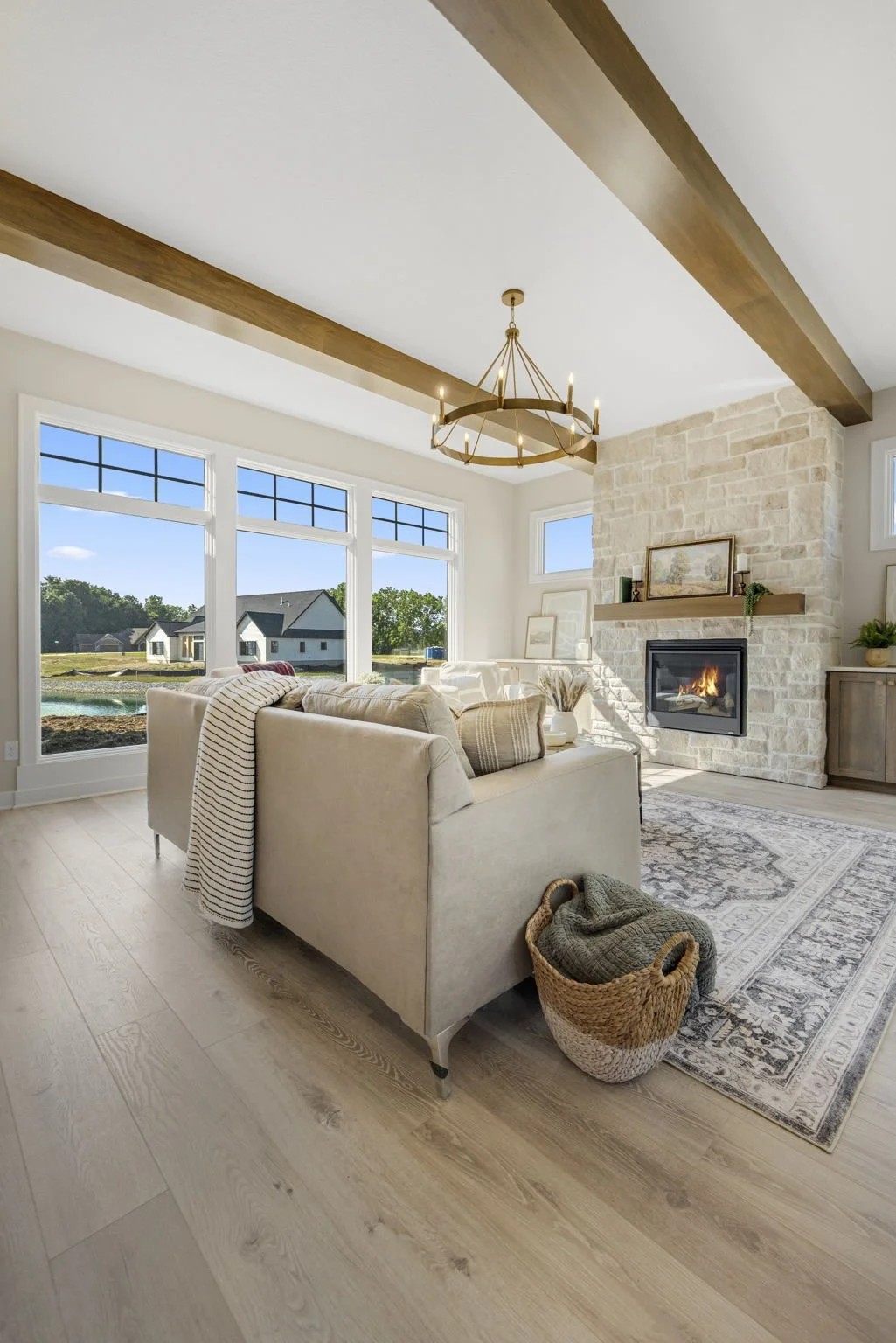 A cozy living room with large windows showing a view of a neighbor's house and yard, a beige sofa with striped and solid pillows, a basket with blankets, a fireplace, and a chandelier hanging from the ceiling.