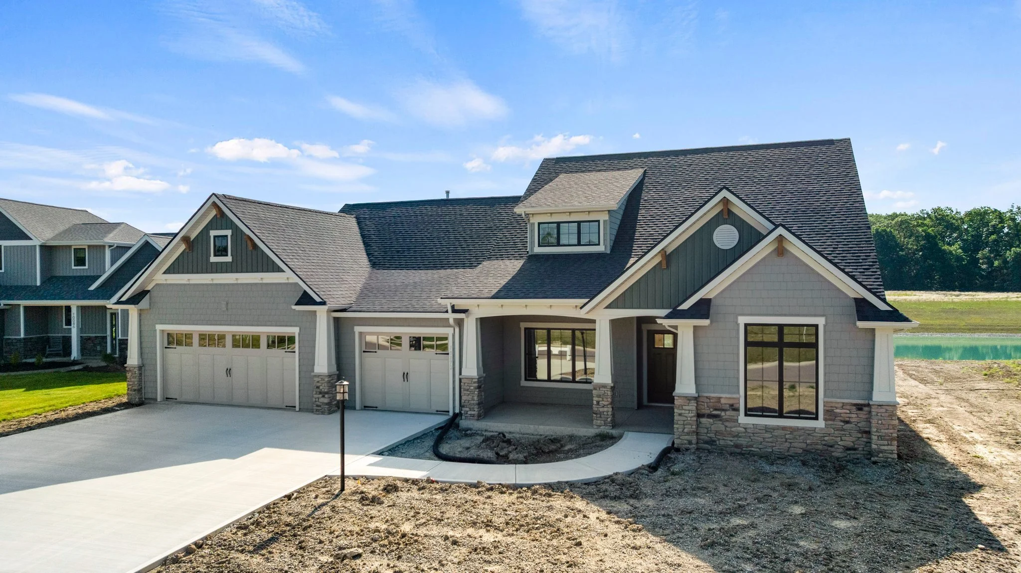 Newly constructed modern house with gray siding, stone accents, three-car garage, and front porch, under blue sky with a few clouds.