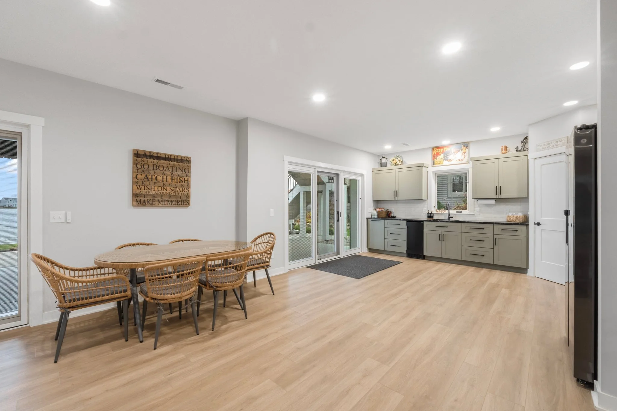 Open kitchen and dining area with light wood flooring, beige cabinets, a sliding glass door and a window, with a sign on the wall reading "LAKE RULES."
