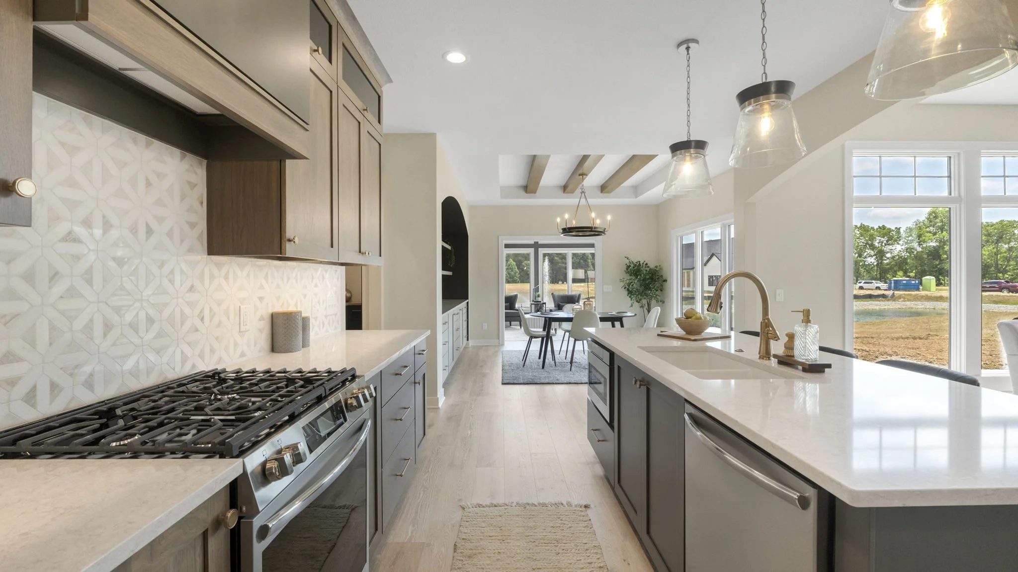 Modern open-concept kitchen with light wooden cabinets, white countertops, and a white island with a sink. Large windows let in natural light, and a view of greenery outside. The kitchen space flows into a dining and living area with a chandelier ove