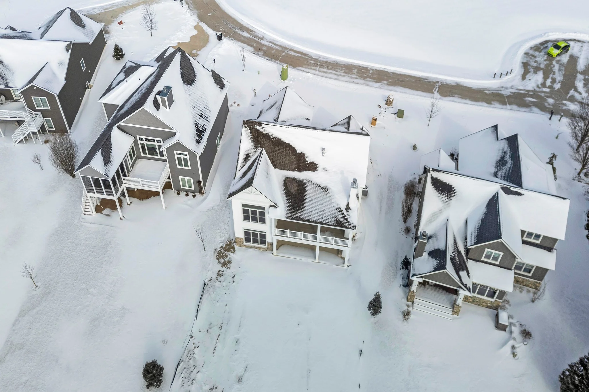 Aerial view of four houses with snow-covered roofs in a neighborhood, showing snow-covered yards and a street with a snow-covered curve and a parked green car.