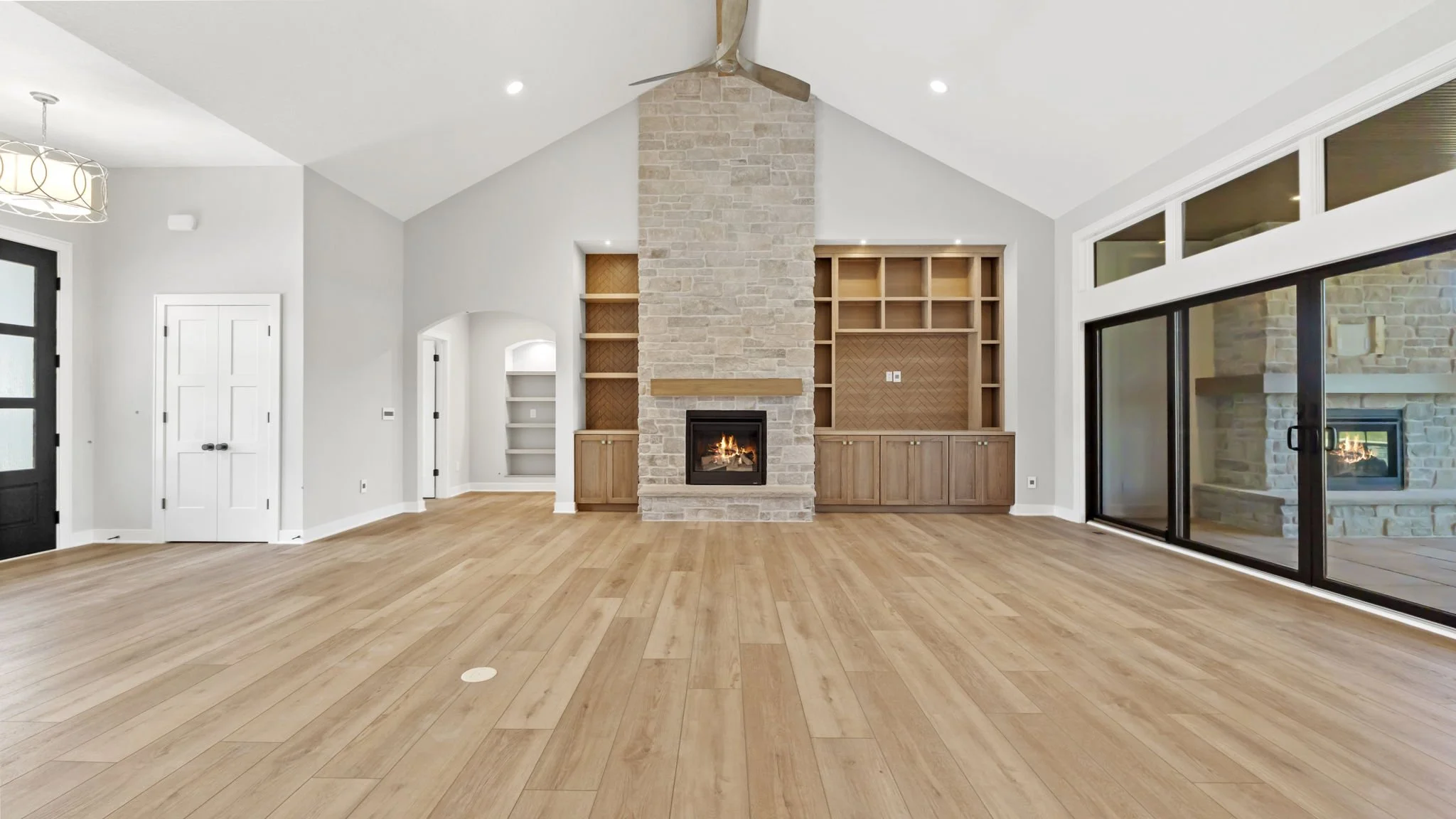 Living room with light wood flooring, a central stone fireplace with a wooden mantel, built-in shelves on either side, and large sliding glass doors leading outside.