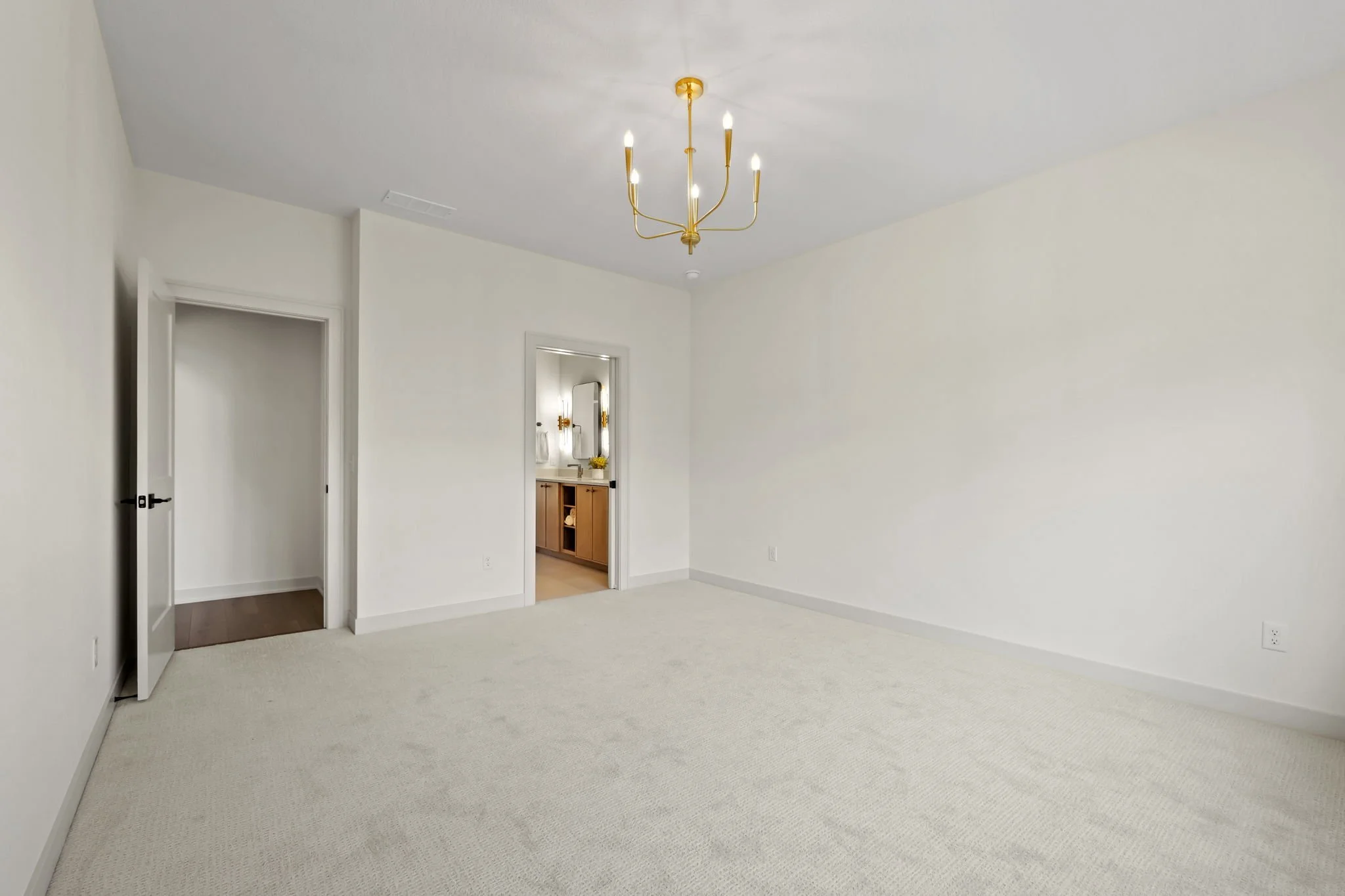 Empty bedroom with white walls, beige carpet, gold chandelier, open closet, and small bathroom with wooden vanity.