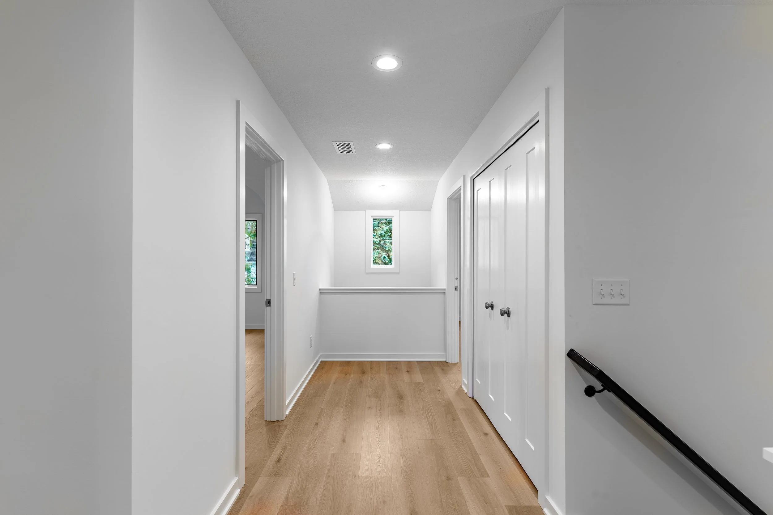 Empty hallway in a modern home with white walls, wooden flooring, ceiling lights, two small windows, and closed closet doors.