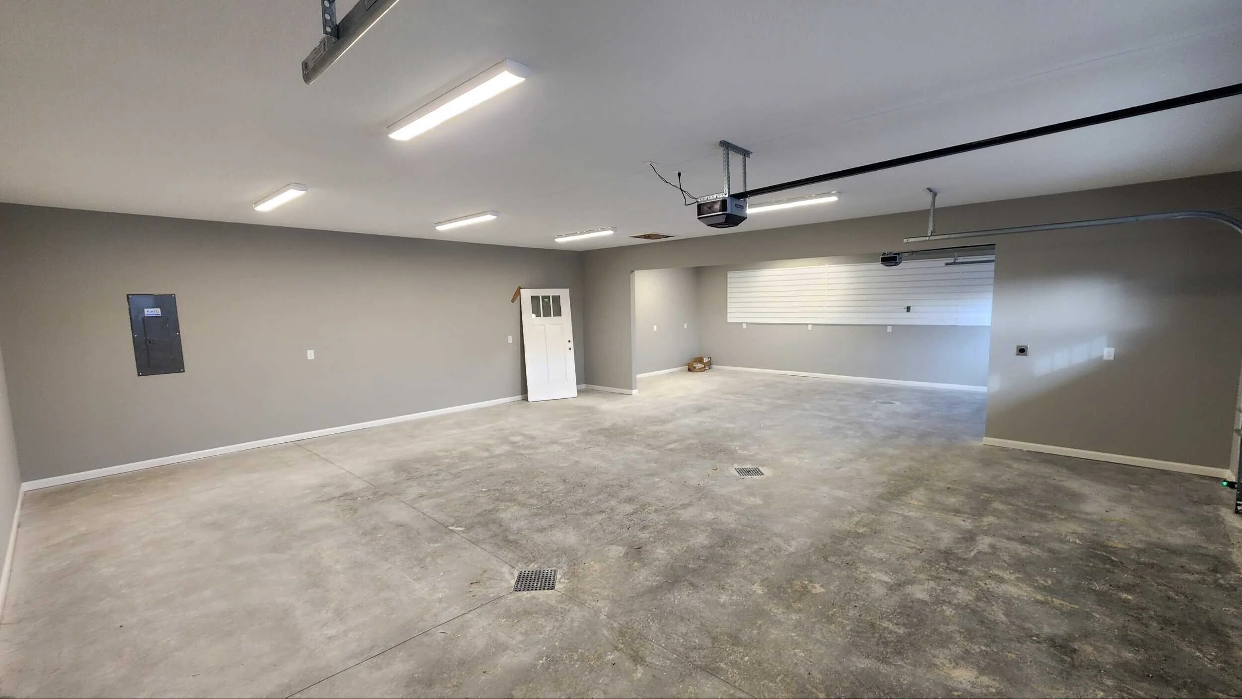 Empty garage with gray walls, concrete floor, fluorescent lighting, garage door openers, and a small window with blinds.