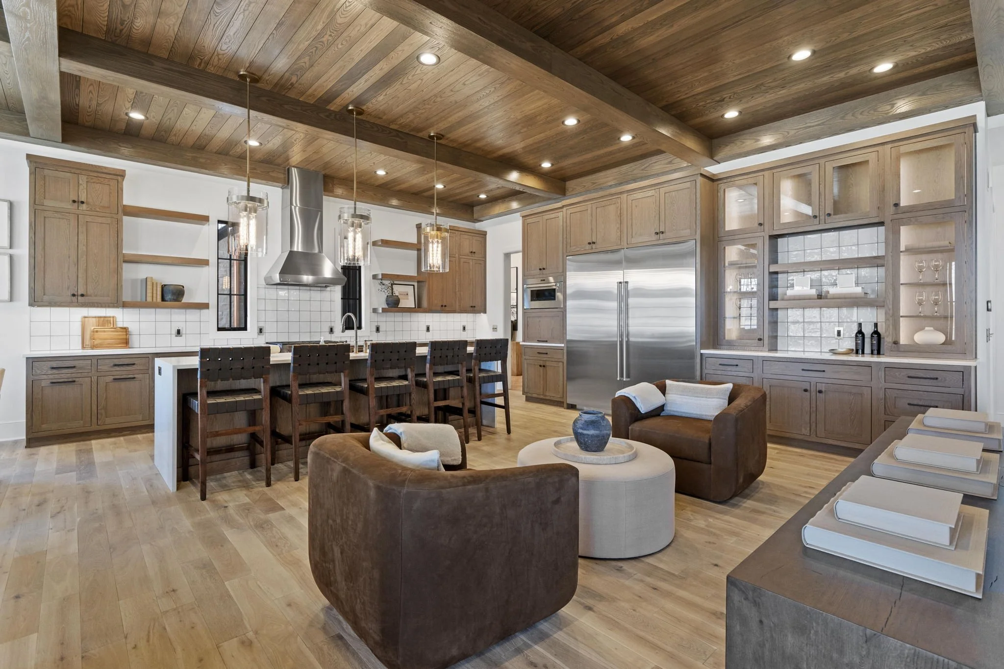 Open-concept kitchen and living room with wooden cabinetry, a large stainless steel refrigerator, a kitchen island with bar stools, a wood-paneled ceiling with recessed lighting, and cozy seating area with two brown armchairs and a round ottoman.