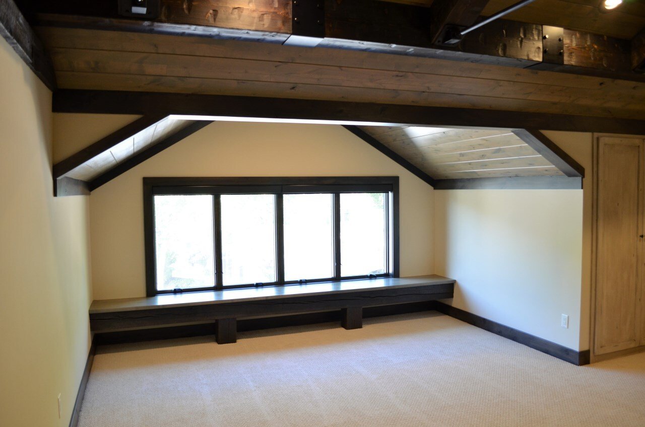 Empty attic room with sloped ceiling, large window with a built-in bench, wood paneling, and beige carpeting.