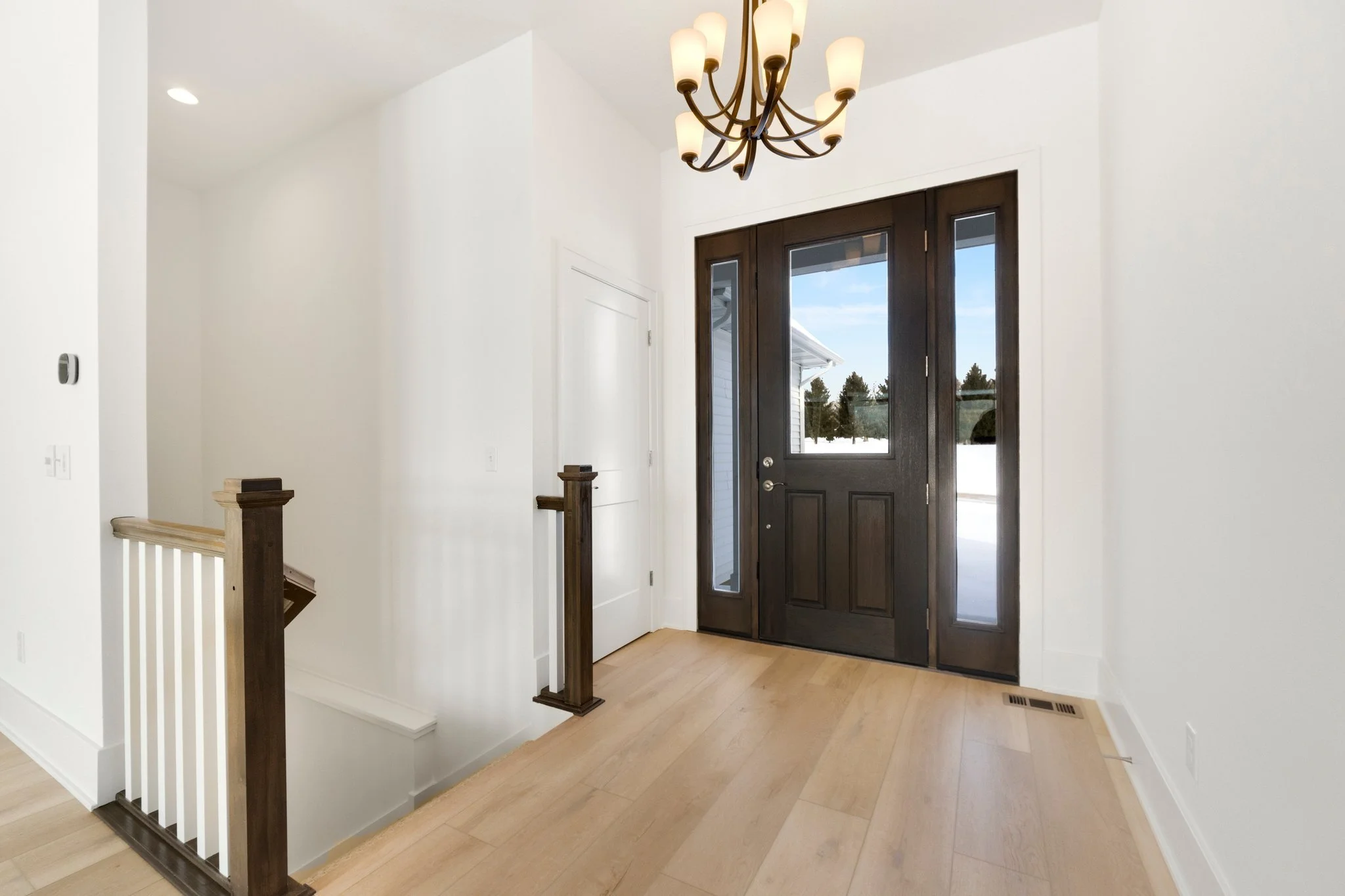 Entryway with dark front door, side windows, chandelier, wooden staircase railing, white walls, light wood flooring, vent on the floor.