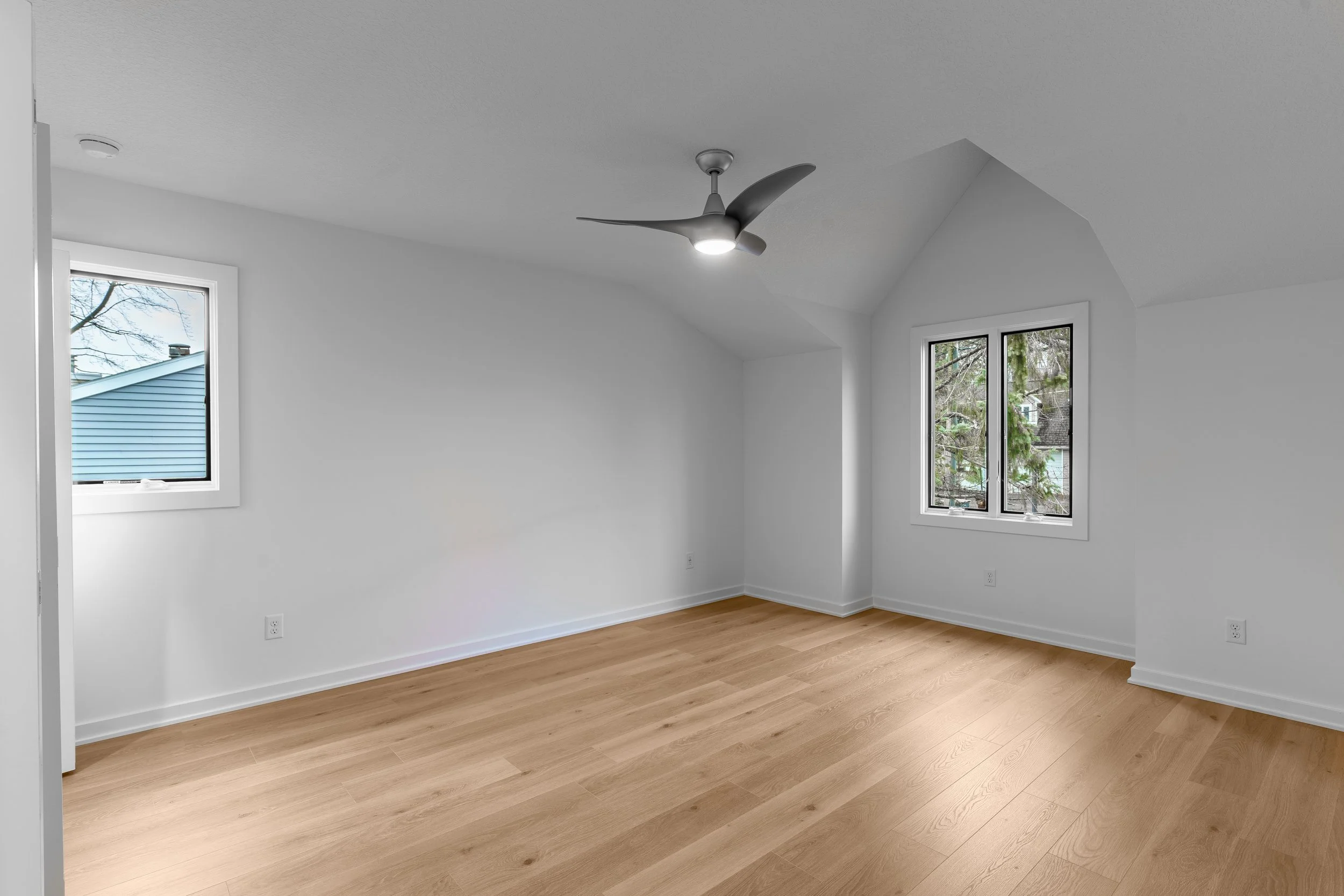 Empty room with white walls, wood flooring, two windows, and a ceiling fan with a light.