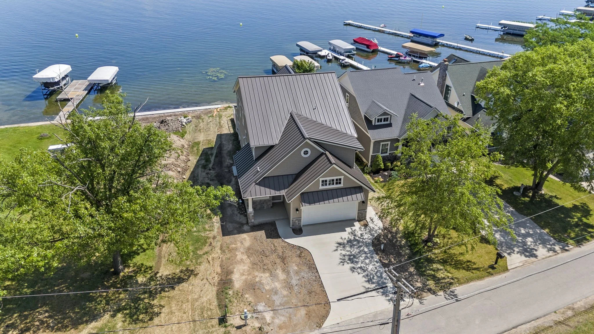 Aerial view of a house near a lake with boat docks and boats, surrounded by trees and neighboring houses.