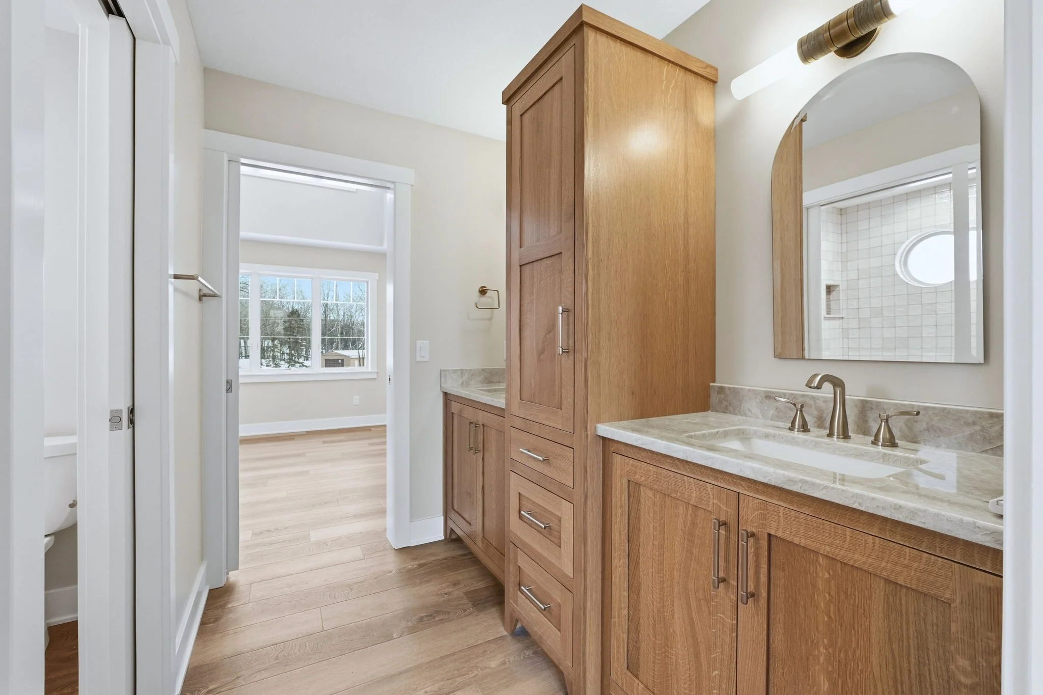 Bathroom with a wooden vanity, marble countertop, and a sink with a brass faucet. Mirror above the sink, wooden cabinetry, and a window with trees visible outside.