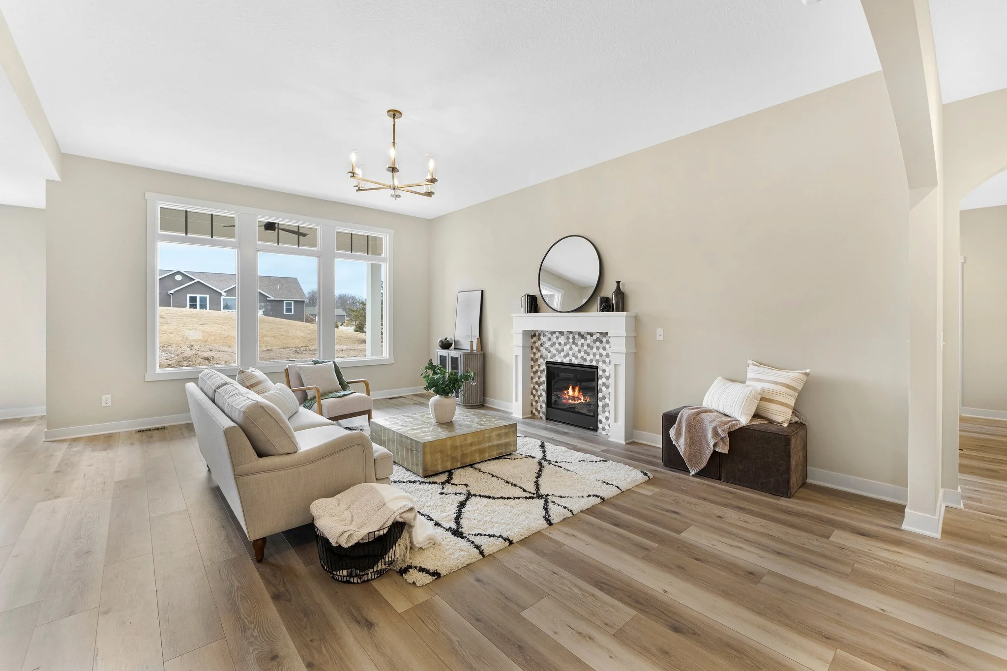 Living room with beige sofa, chair, black and white rug, fireplace, wall mirror, large window, and hardwood floors.