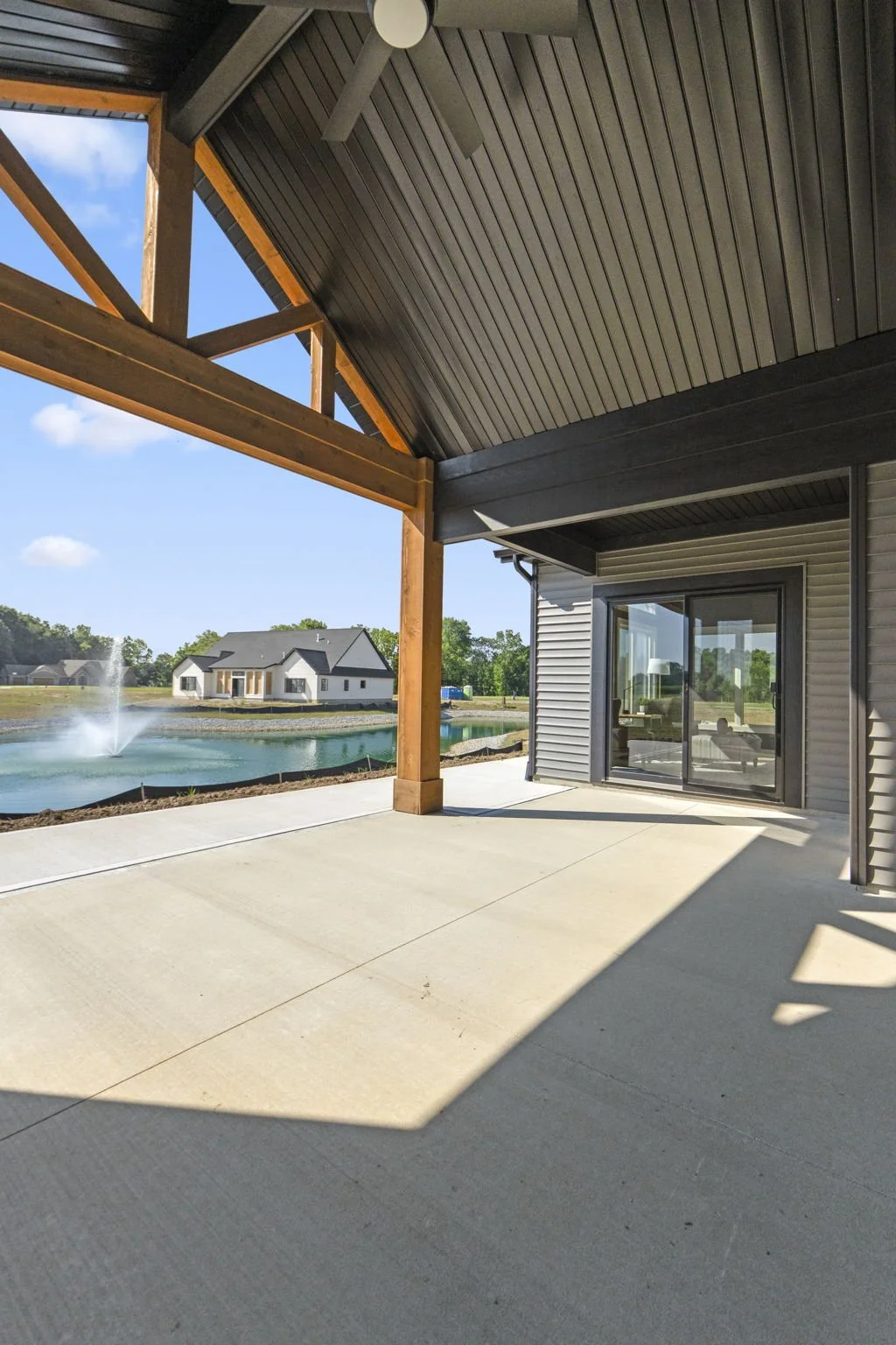 View of a backyard patio with a concrete floor, a sliding glass door, and a modern house with a pond and fountain in the background.