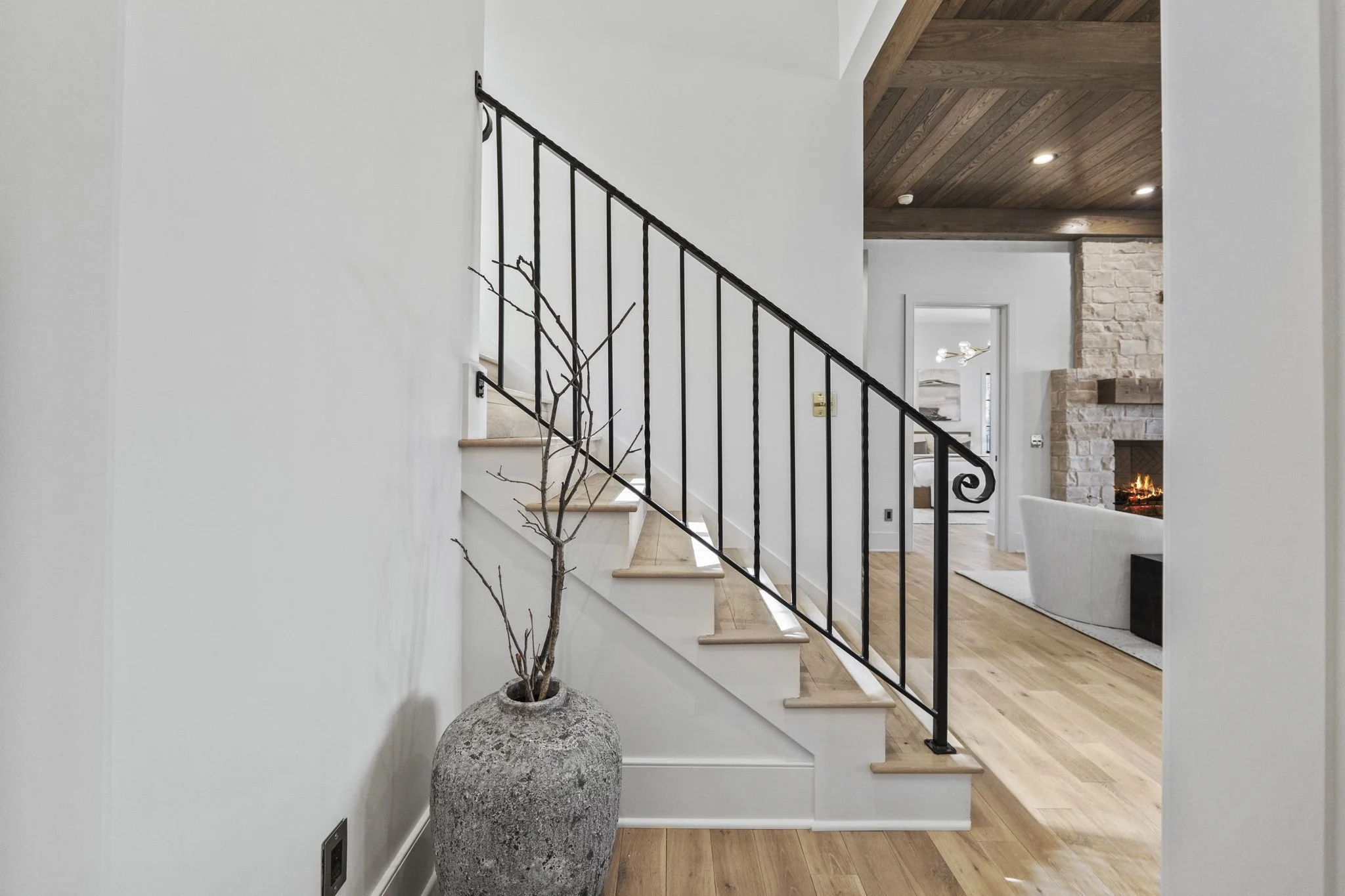 Interior view of a modern house with a staircase, a bare branch in a large textured pot, and a living room with a fireplace and wooden ceiling.