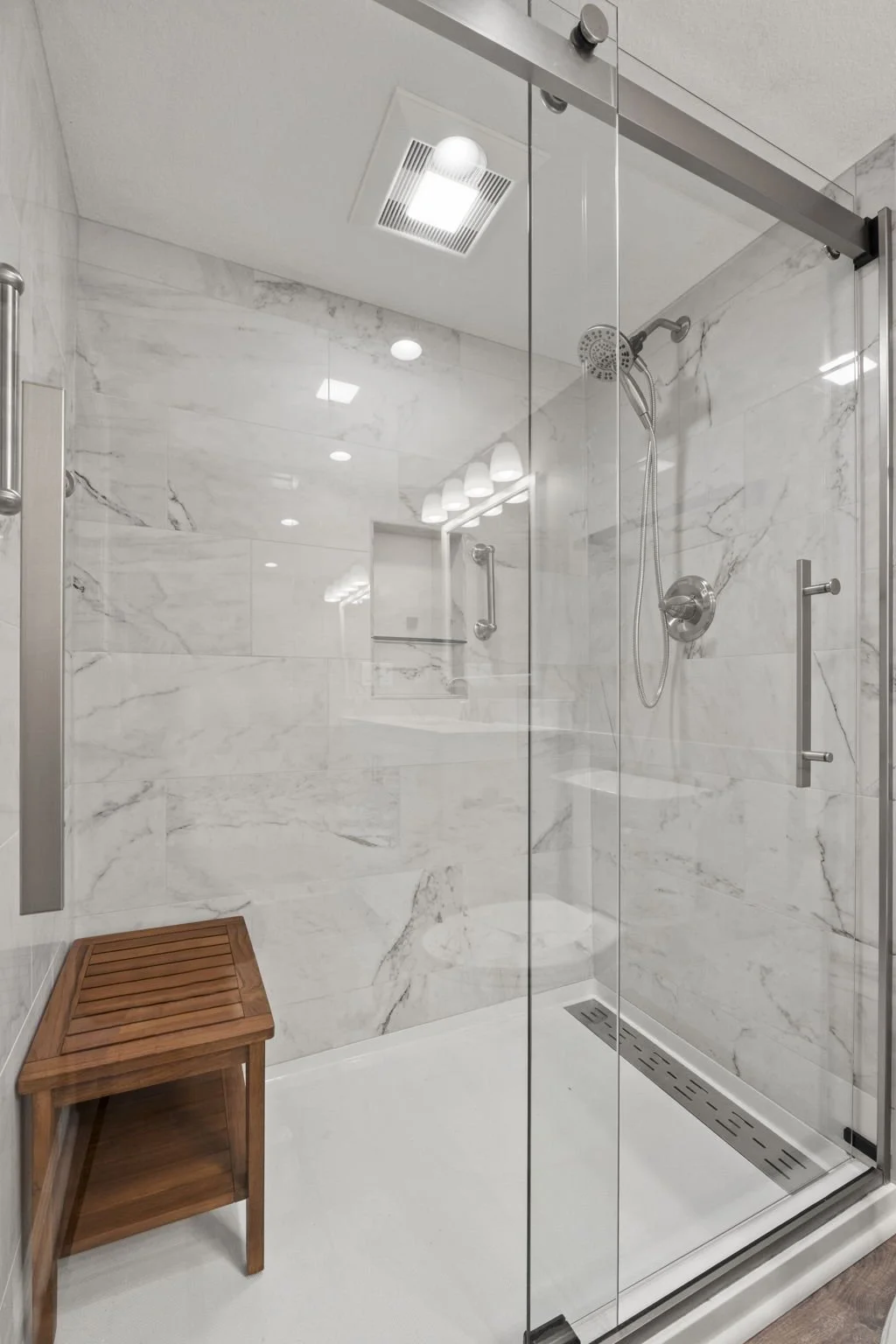 Glass shower enclosure with marble walls, a handheld shower head, a small built-in shelf, and a wooden stool, with ceiling lighting and a vent.