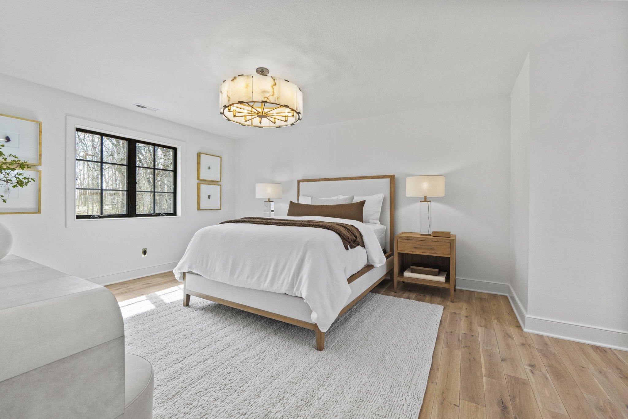 Minimalist bedroom with white walls, wooden furniture, large window showing trees outside, and beige carpet.