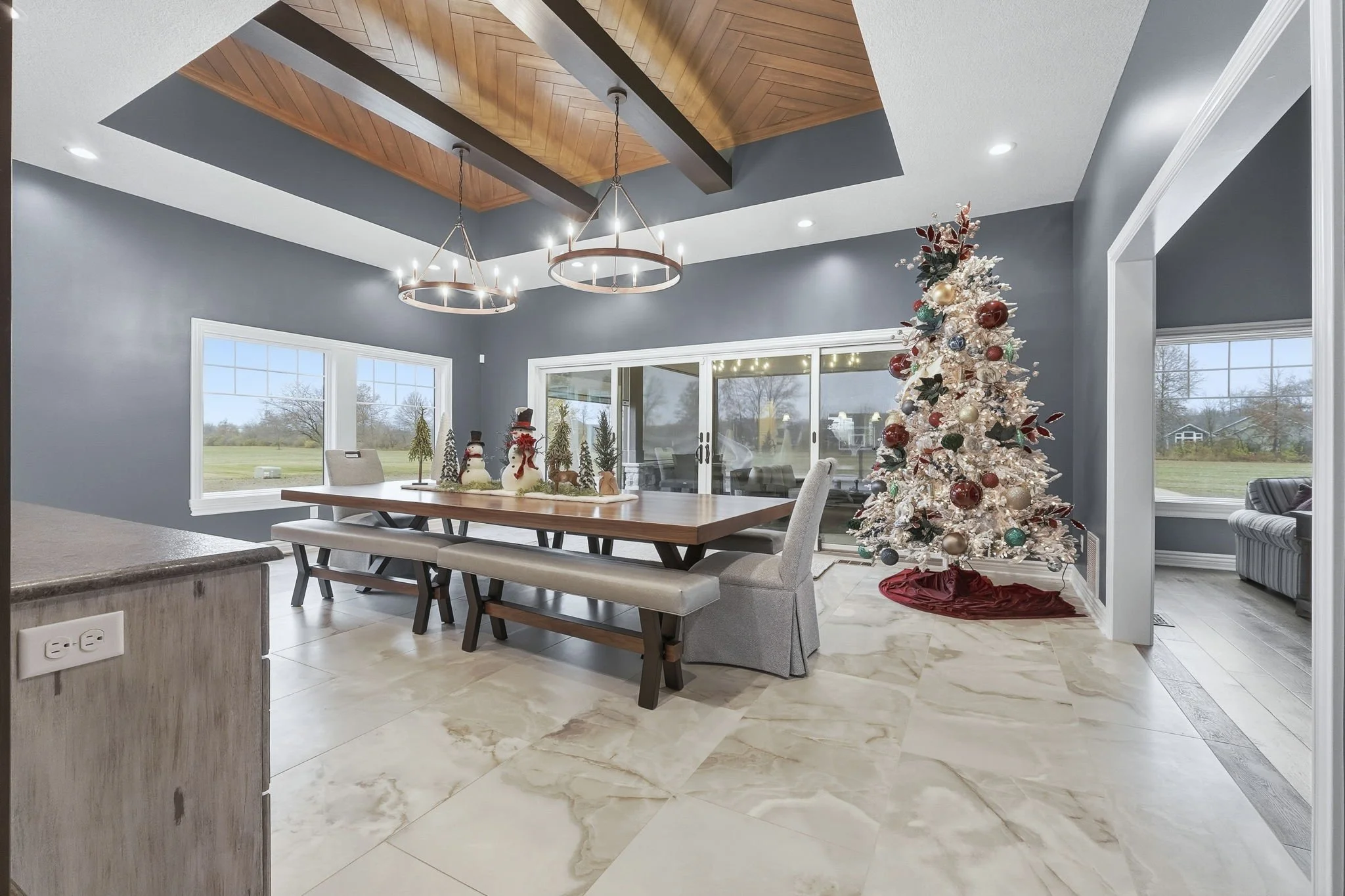 A decorated Christmas tree with ornaments and a red tree skirt in a dining room with gray walls, a wooden ceiling accent, hanging chandeliers, large windows, and holiday decorations on the table.