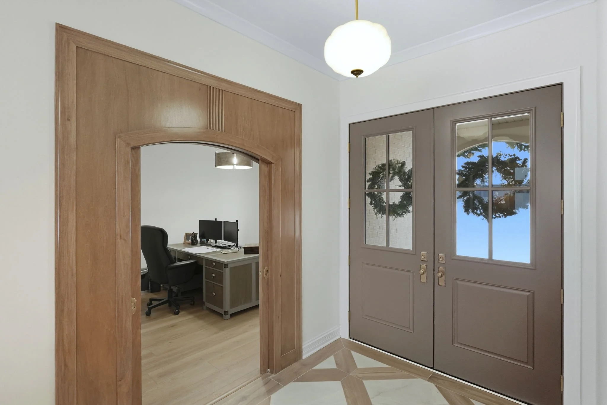 View of an entryway with gray double doors with glass panes, gold hardware, and a wreath, adjacent to a wall opening leading to an office with a black desk, office chair, and computer.