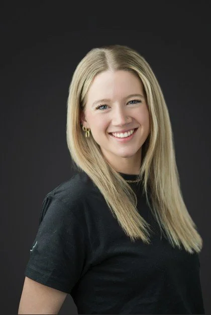 A young woman with long blonde hair smiling, wearing a black t-shirt and gold hoop earrings, posed against a dark background.