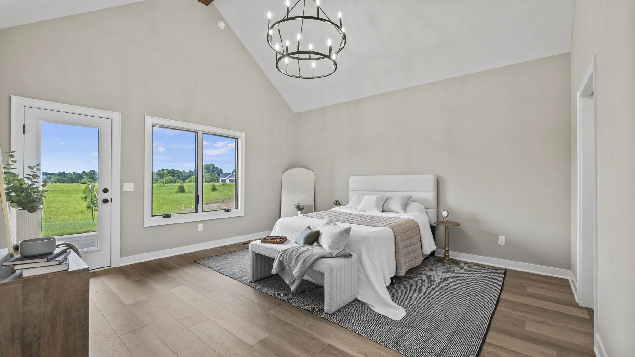 A bedroom with a sloped ceiling, a large bed with white bedding, a bench at the foot of the bed, and a large window showing green fields outside.