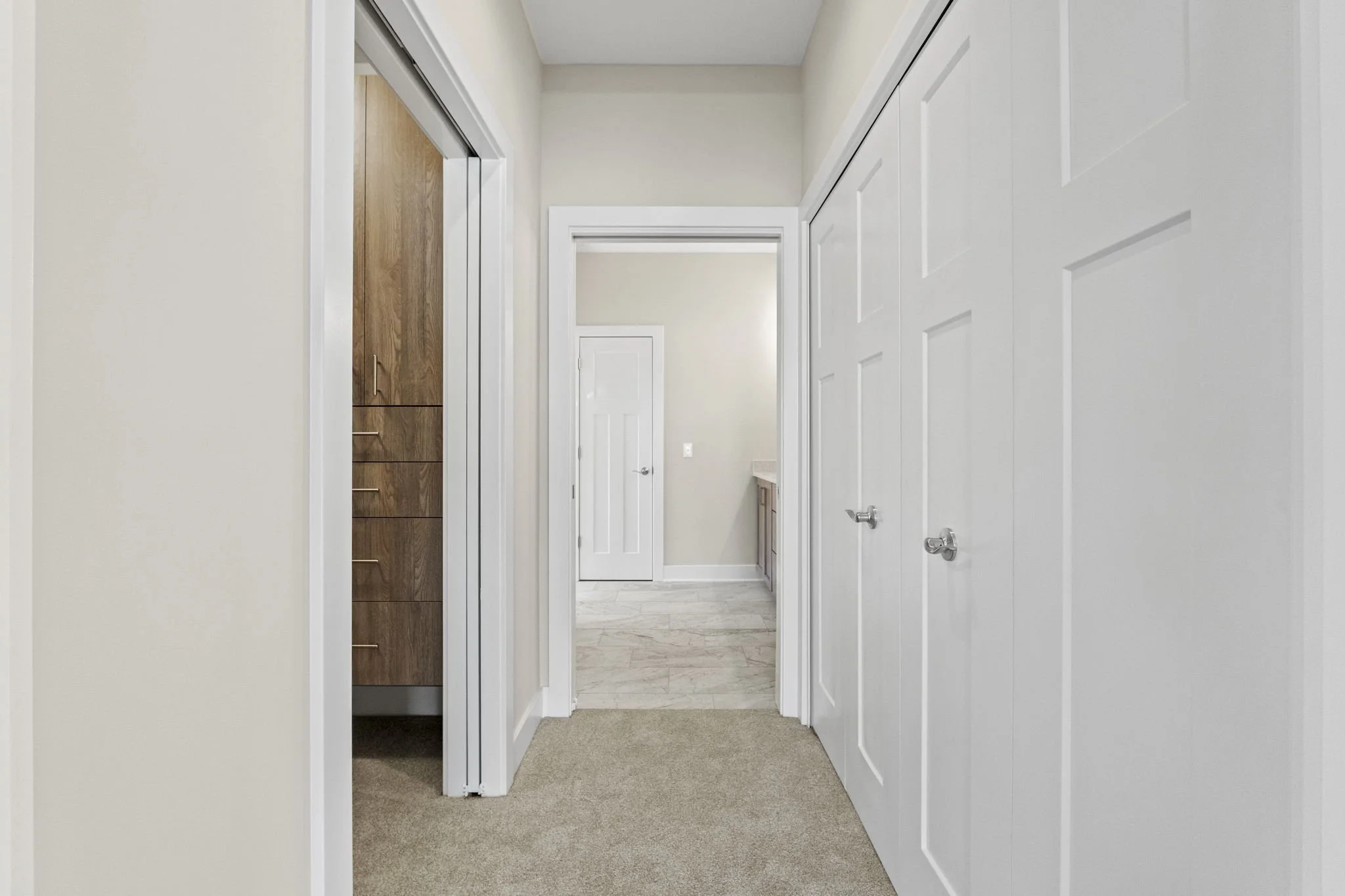 View of an interior hallway with beige walls, white doors, and carpeted flooring leading to a bathroom with tile flooring and wooden cabinets.
