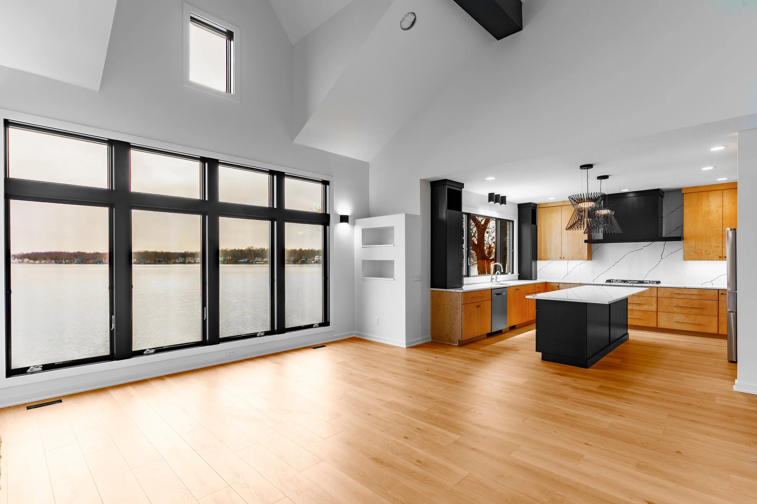 Modern kitchen and living area with large windows overlooking a lake, featuring light wood floors, white walls, and black and wood cabinetry.