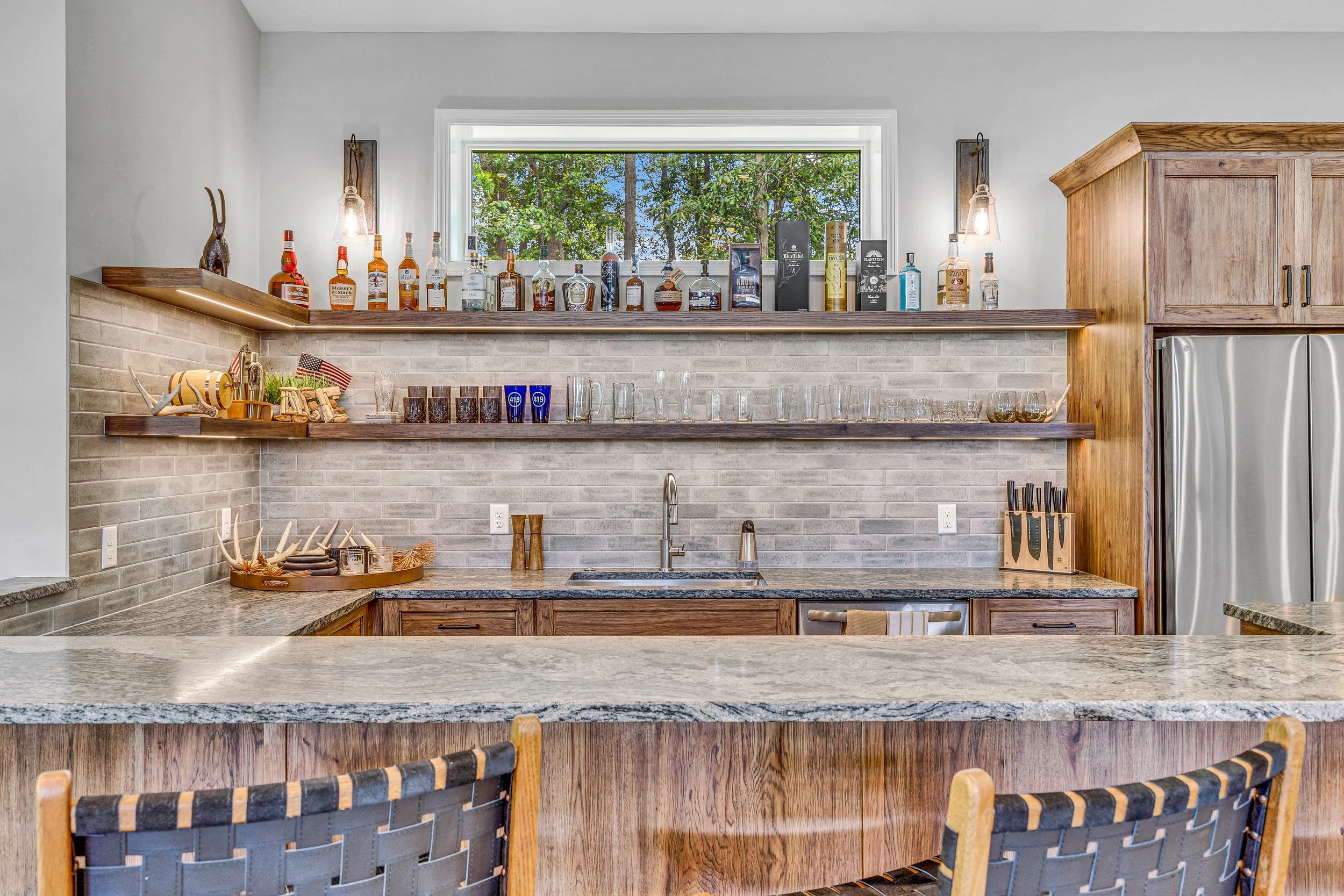 Kitchen with open shelves displaying bottles of liquor, glasses, and kitchen utensils, a window with trees outside, a granite countertop, and wooden cabinets.
