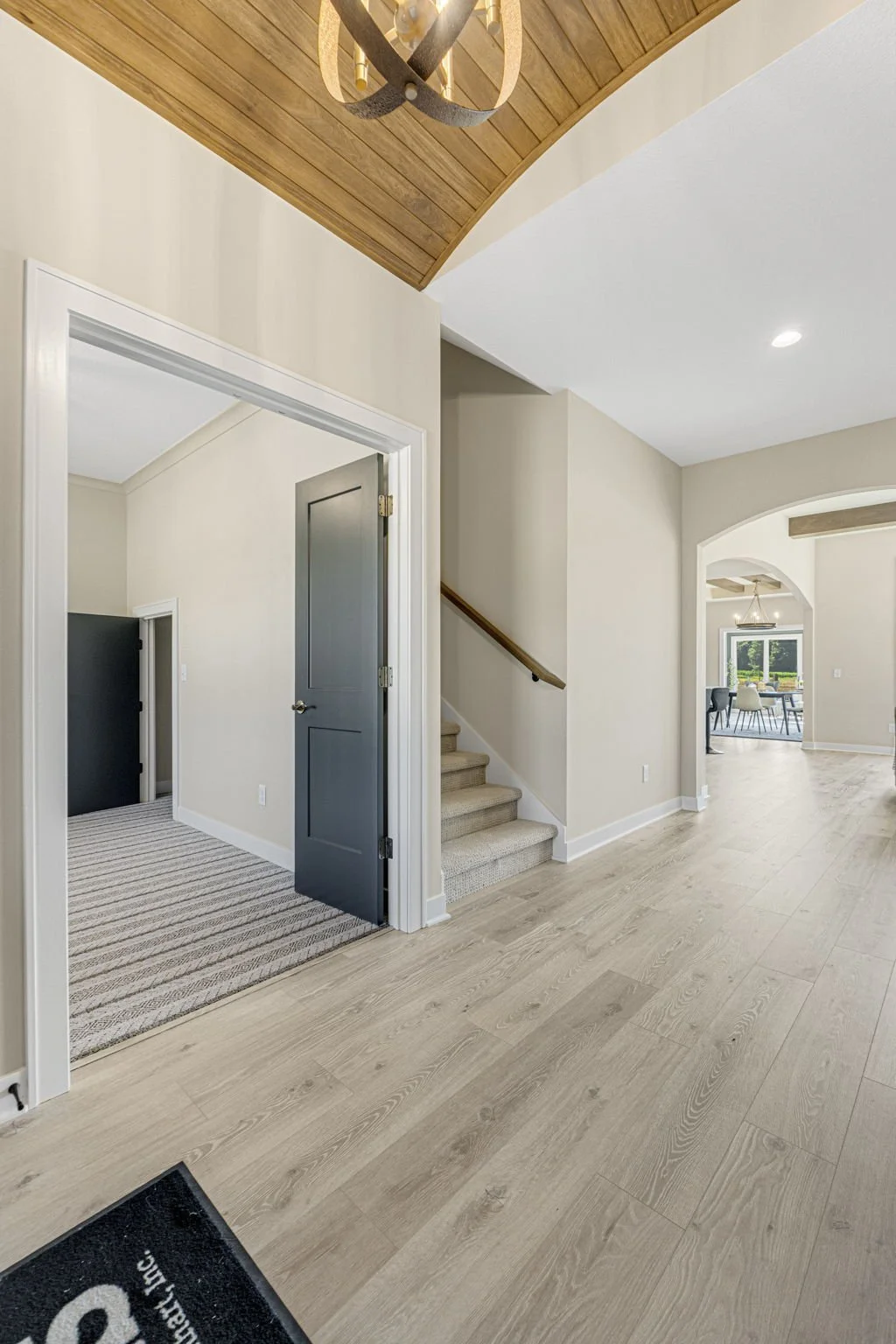 Interior view of a modern home with light wood flooring, beige walls, and a staircase with carpeted steps and a wooden handrail. There is a doorway to a room with a gray door and a carpeted floor. In the background, there's a dining area with a table