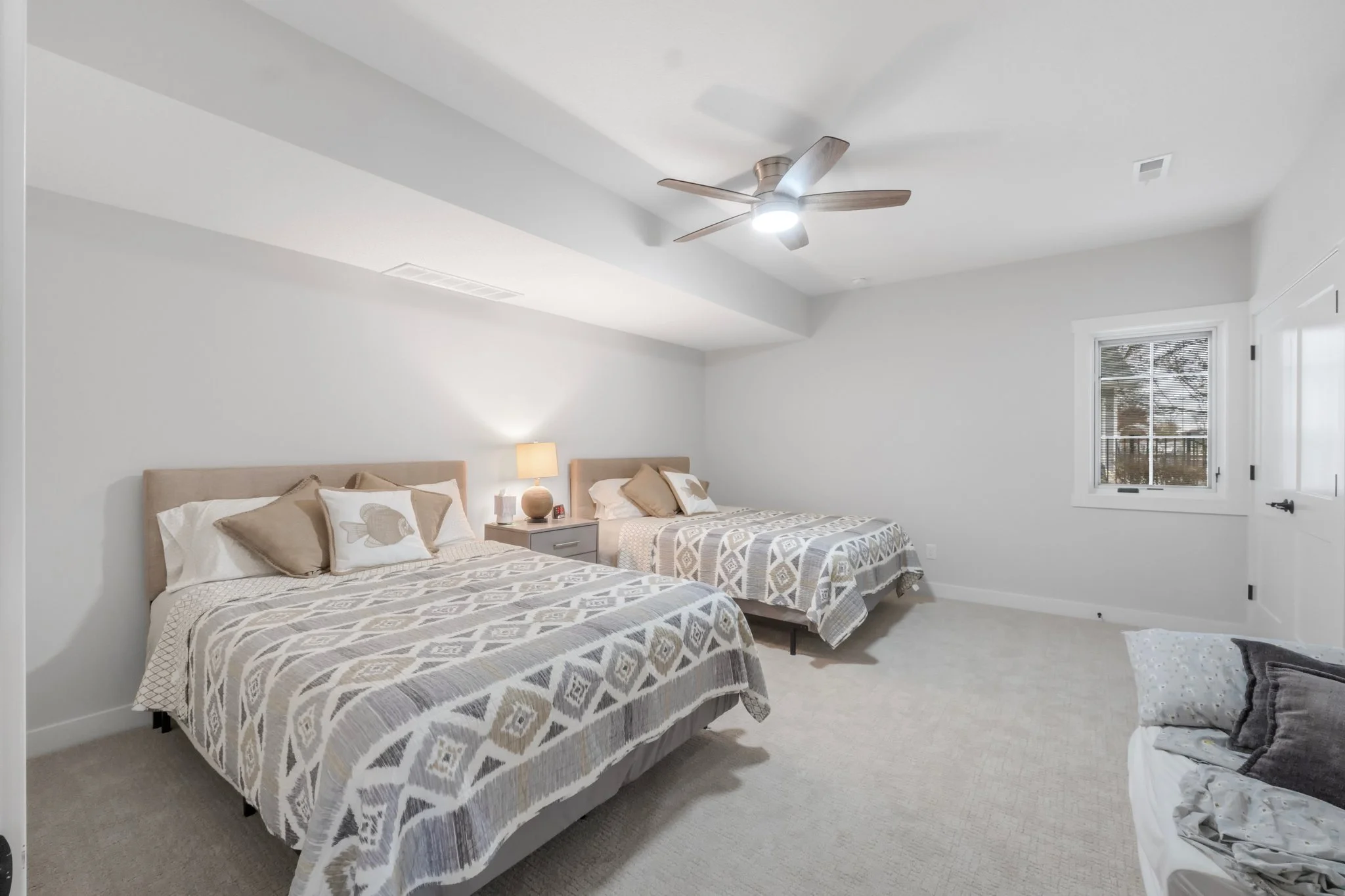 A bedroom with two beds, beige headboards, matching patterned bedspreads, a nightstand with a lamp between the beds, and a window with blinds, in a minimalist style.