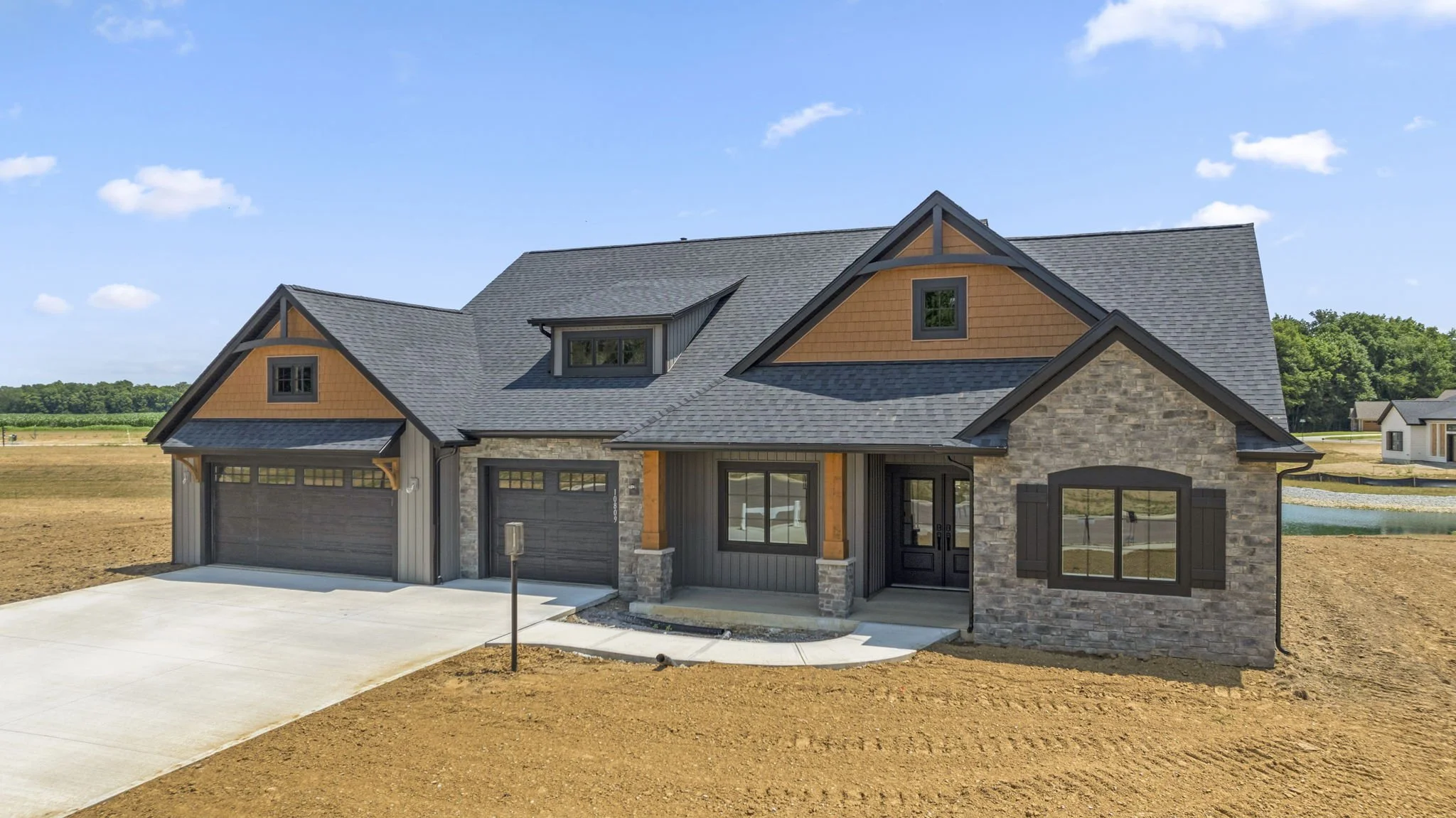 New modern house with gray brick exterior, wooden accents, and a two-car garage, situated on a dirt lot in a suburban area under a partly cloudy sky.