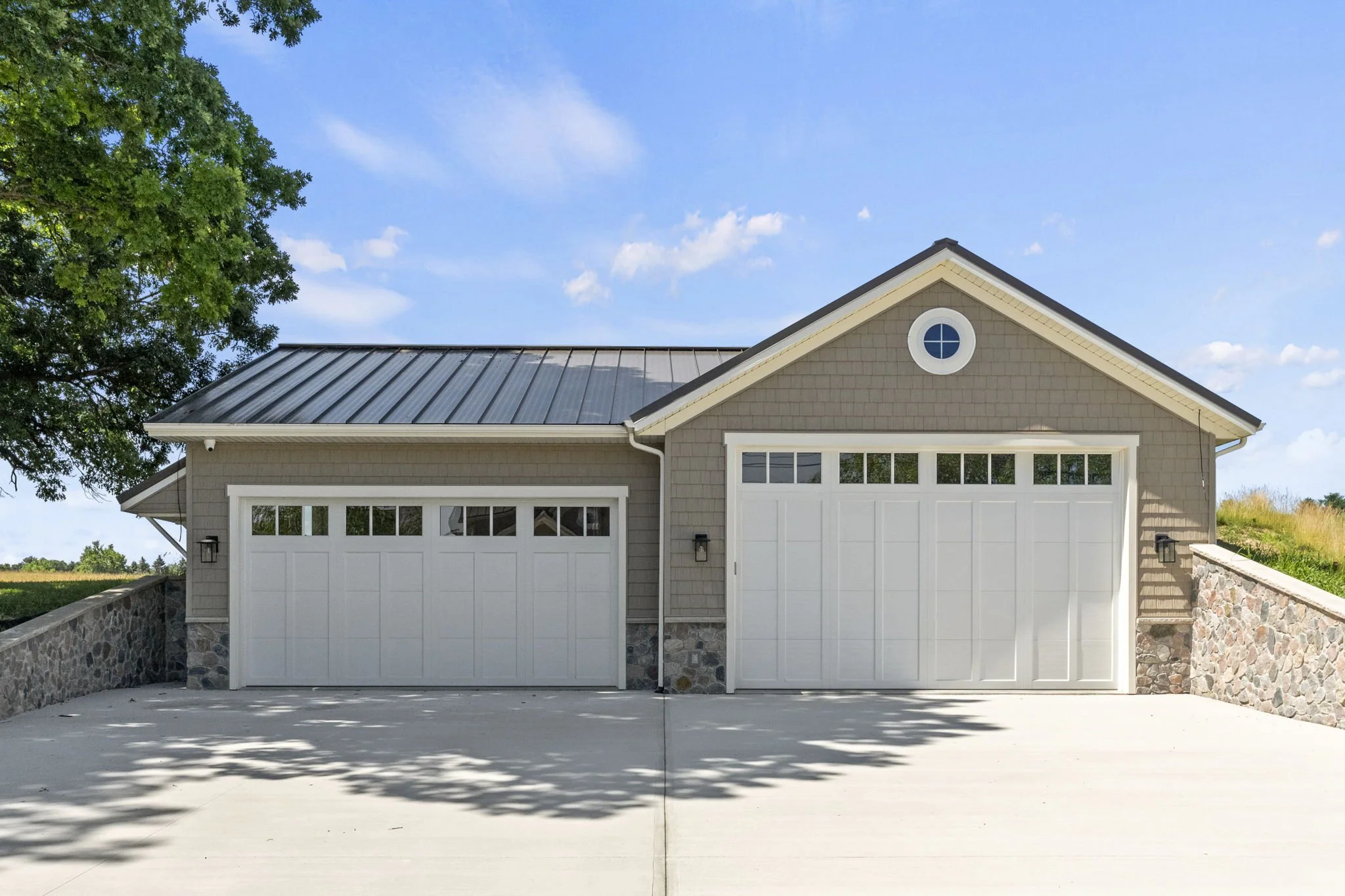 A modern two-car garage with beige siding, white garage doors, a stone foundation, and a metal roof; surrounded by a concrete driveway with shadows of trees, and a clear blue sky with some clouds.