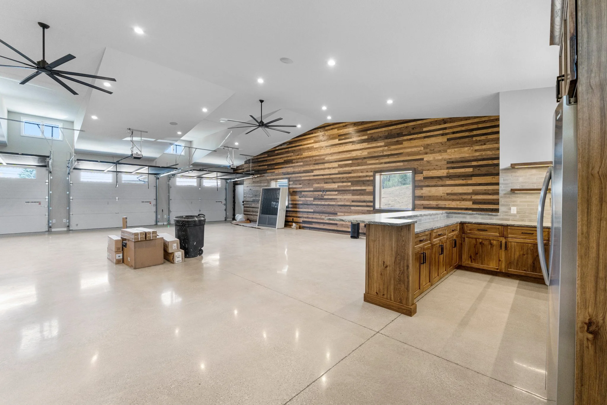 Spacious garage with a wooden accent wall, kitchen cabinets, and windows, with some boxes and a trash can in the center.