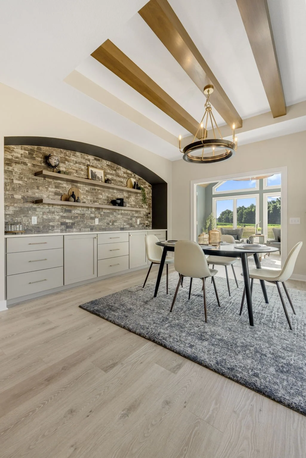 Modern dining room with a round black table, six light-colored chairs, a gray textured rug, a built-in cabinet with shelves against an arched brick wall, large window displaying a green outdoor landscape, and a ceiling with wooden beams and a circula