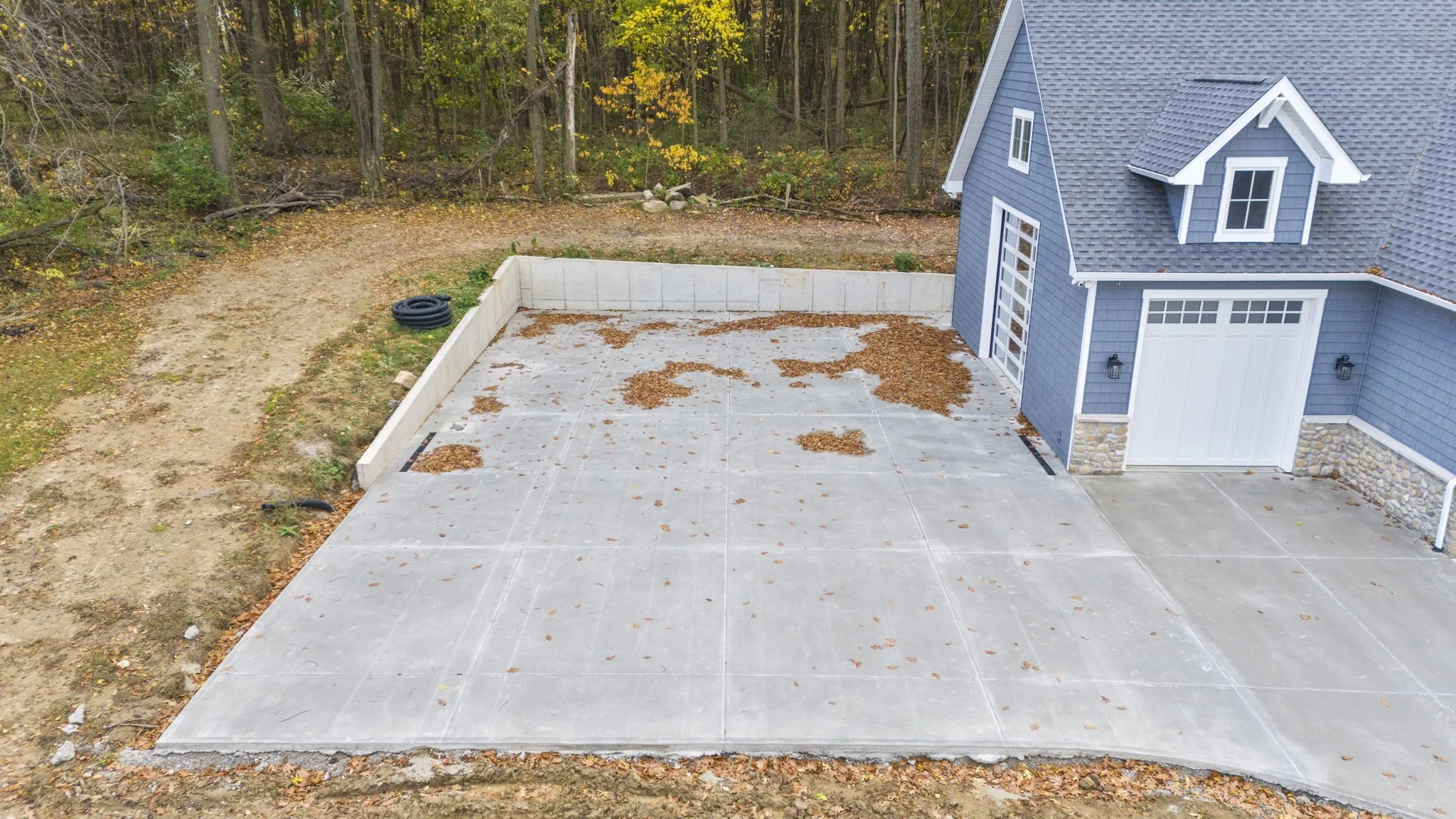 An empty concrete driveway with some fallen leaves, adjacent to a blue house with a garage, stone accents, and surrounded by trees and wooded area.